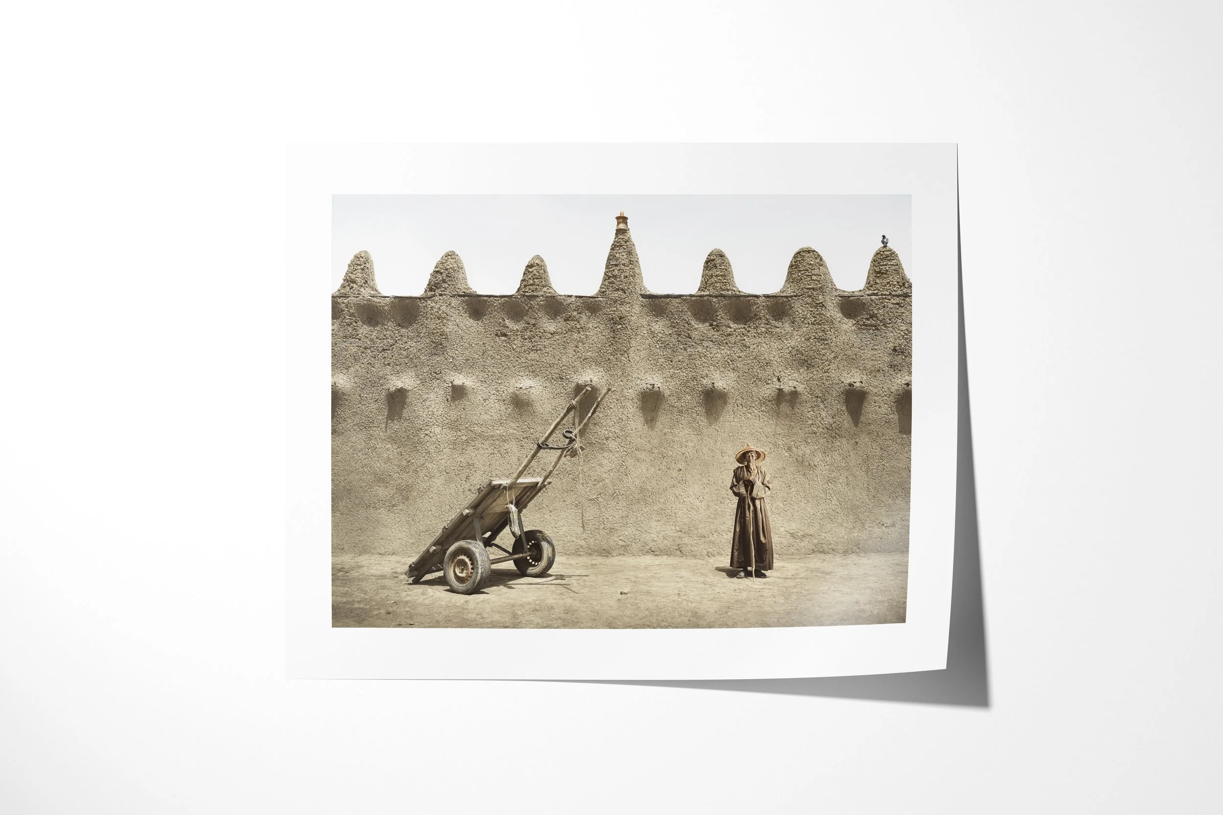 Fine art paper print of Ouman, showing Ouman standing beside a two-wheeled handcart against the sculpted wall of an earthen mosque.