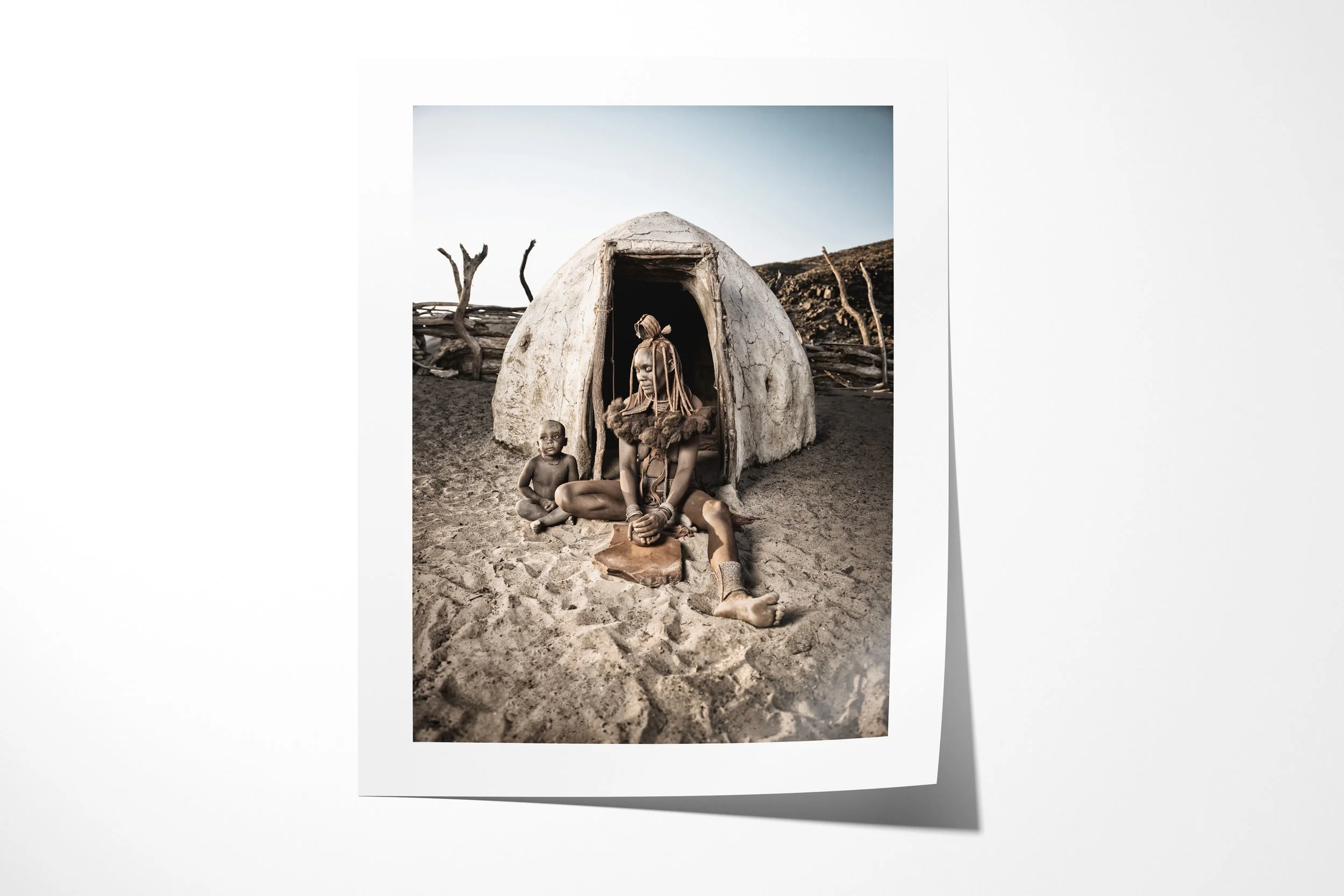 Fine art paper print of Ngaringombe And Her Son, showing Ngaringombe seated with her son outside a domed Himba dwelling while grinding ochre on a stone.