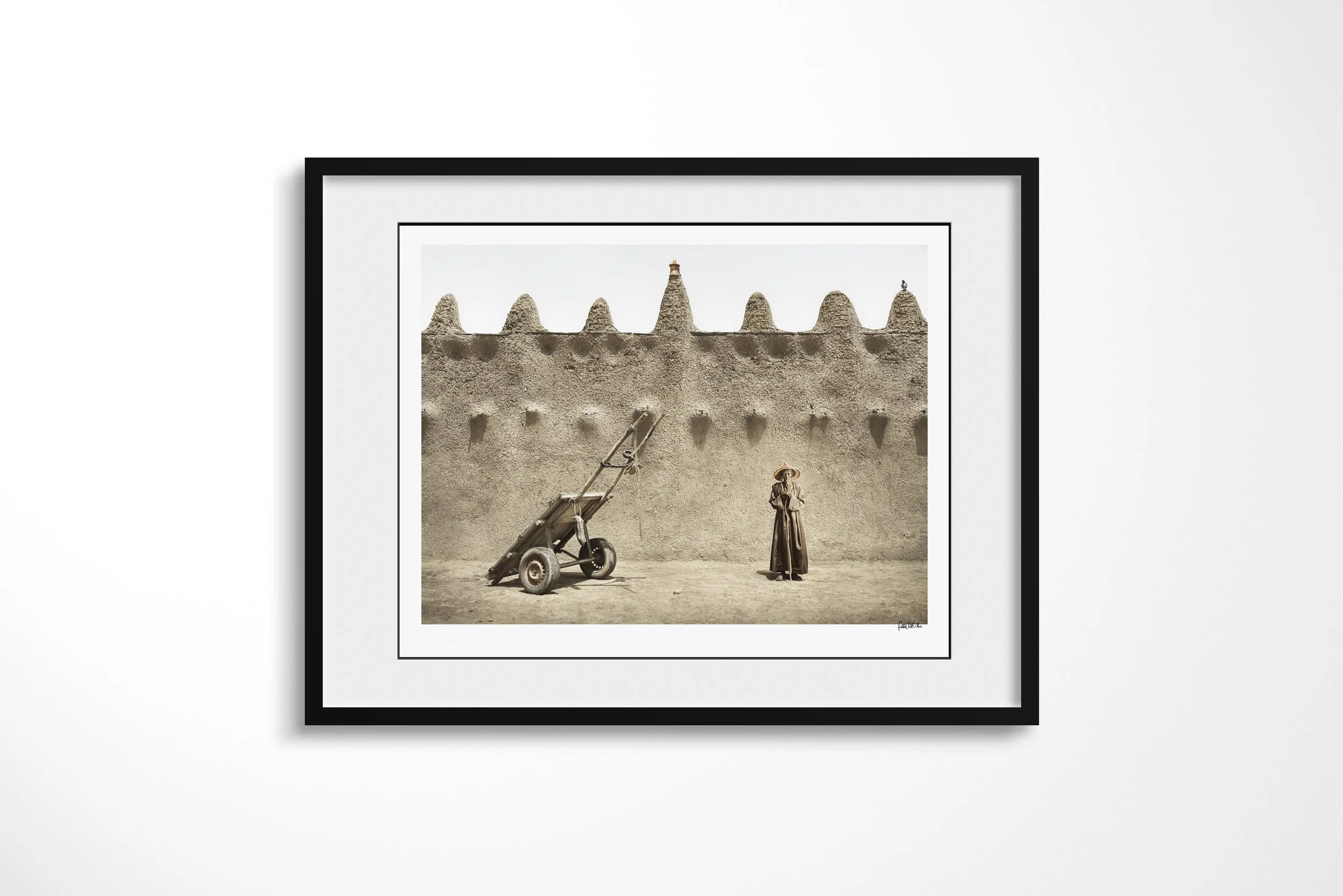 Framed presentation of Ouman in a black frame and white mount, showing Ouman standing beside a two-wheeled handcart against the sculpted wall of an earthen mosque.