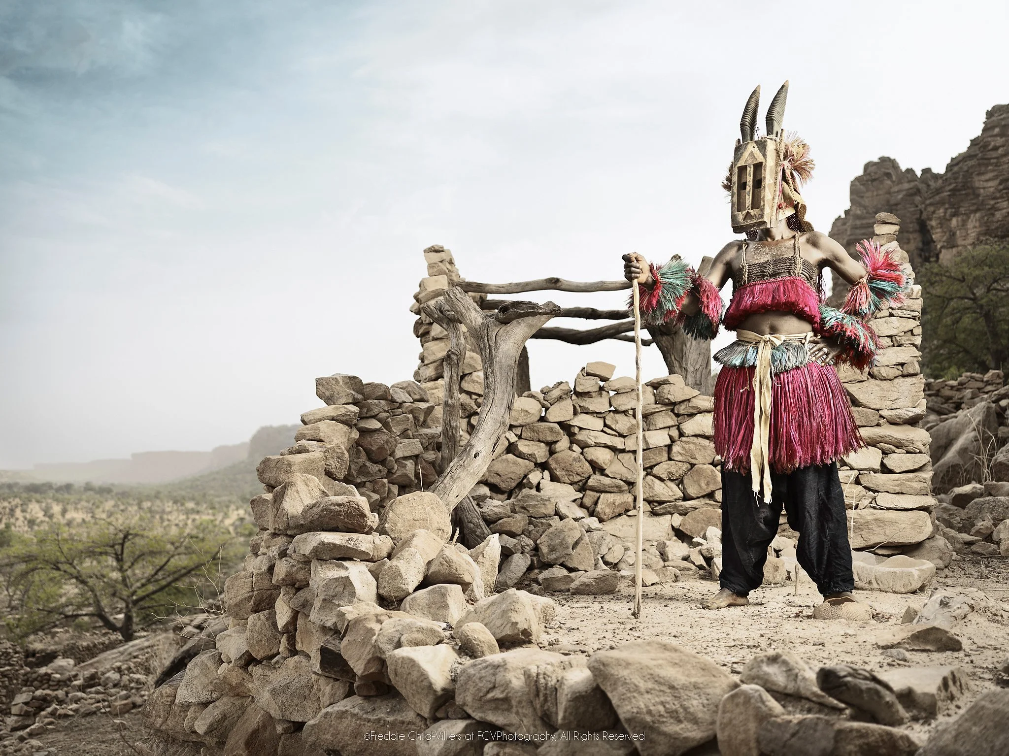 Walu (Mask) II, a Dogon Walu mask holding a staff among the stone walls of a cliff village.