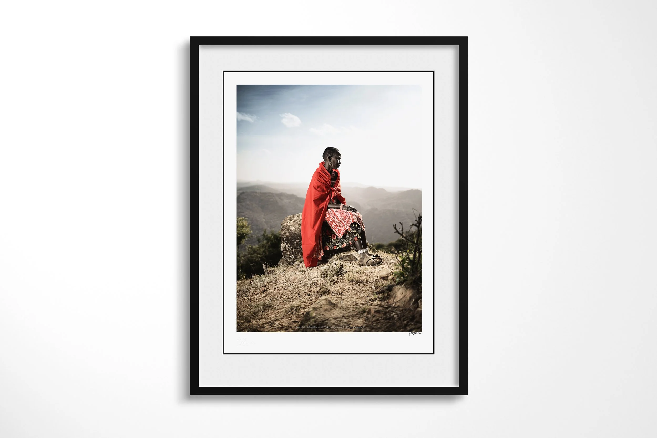 Framed presentation of Msari in a black frame and white mount, showing a Samburu woman in a red wrap seated on a high rock above the Matthews Mountains.