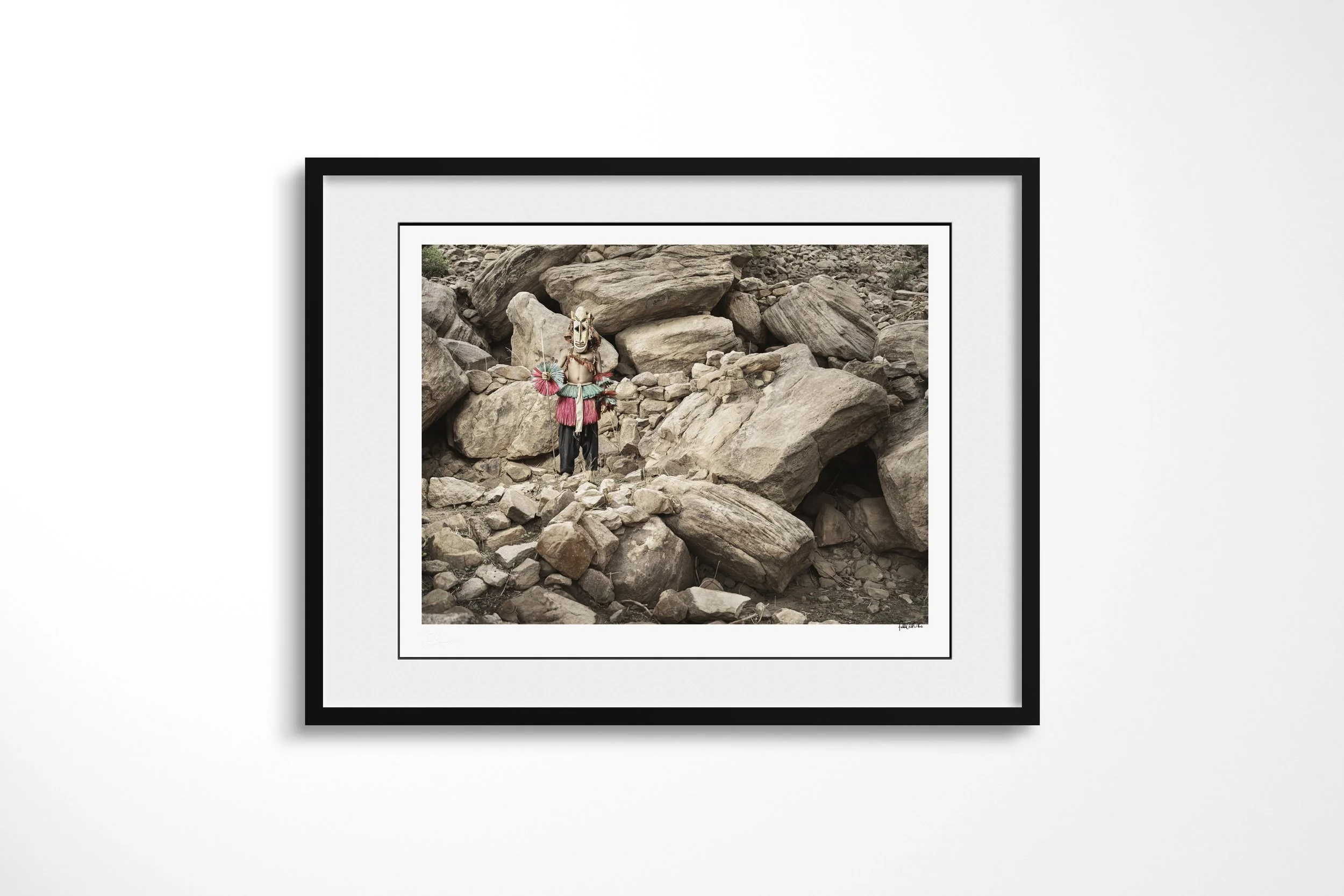 Framed presentation of Djon (Mask) in a black frame and white mount, showing a Dogon antelope mask standing amid collapsed boulders and stone walls.