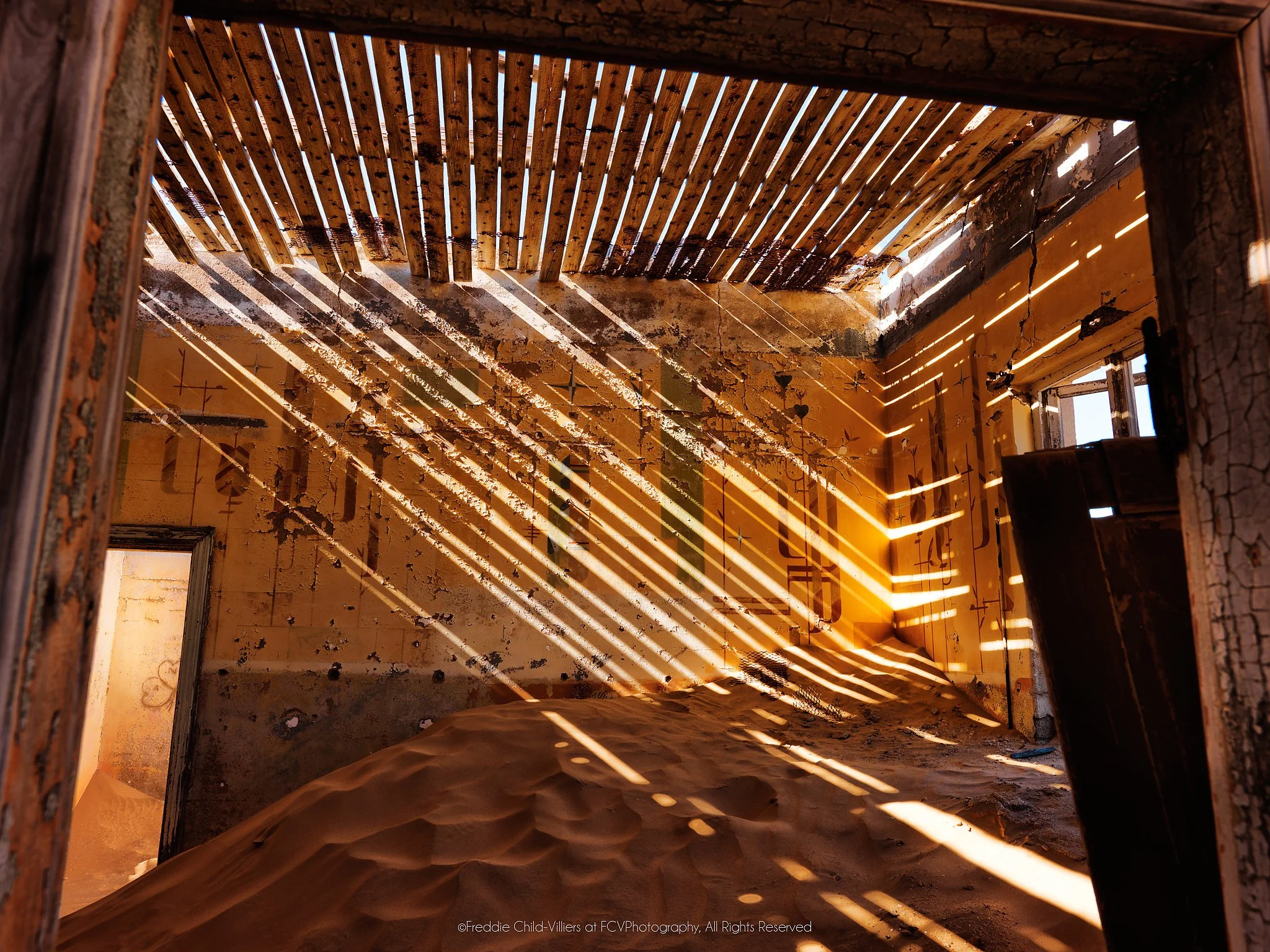 Sand-filled ochre room crossed by diagonal sunlight from a damaged slatted roof, with peeling walls and an open doorway.