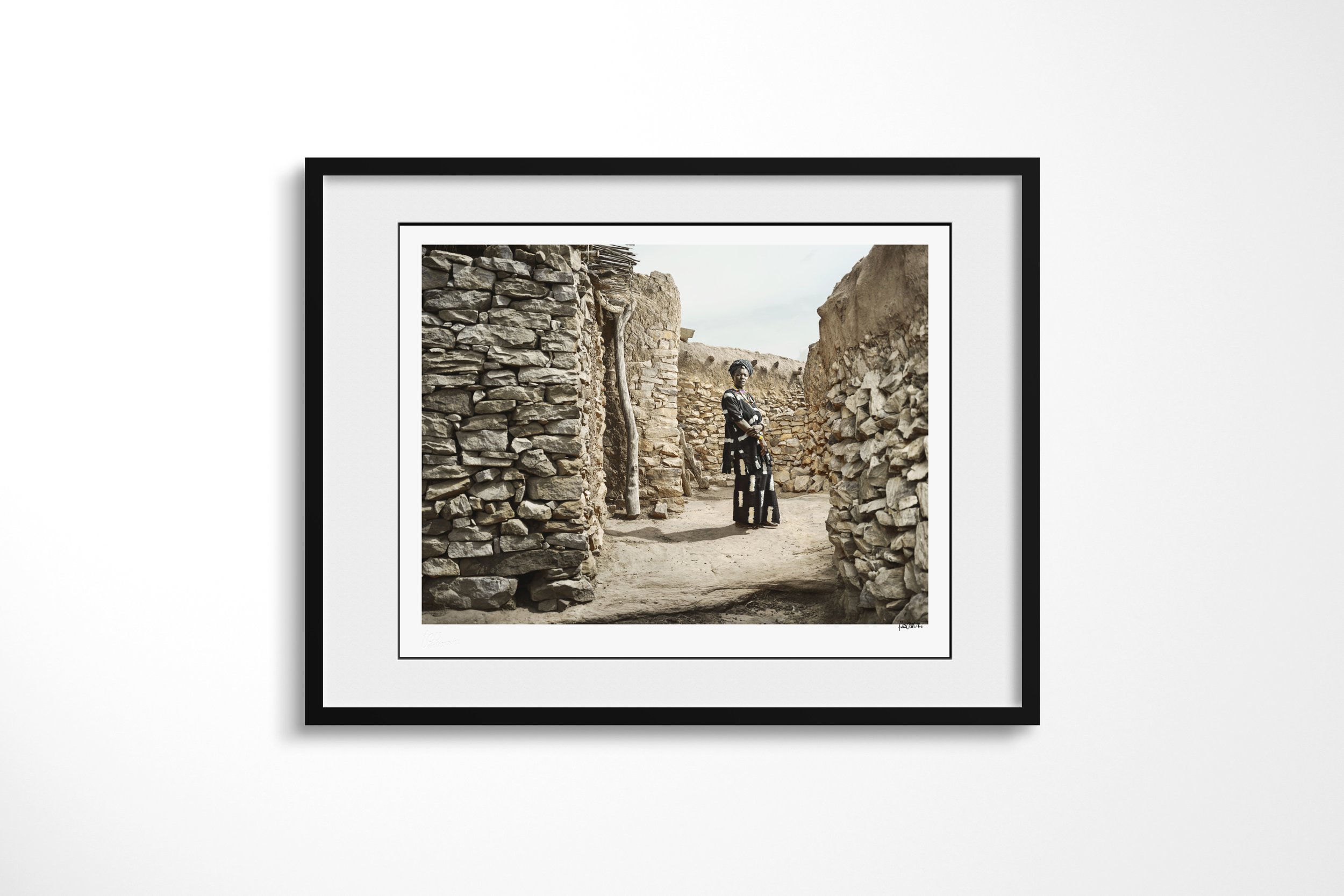 Framed presentation of Yakoto in a black frame and white mount, showing Yakoto standing in a narrow stone passage between the walls of a Dogon village.