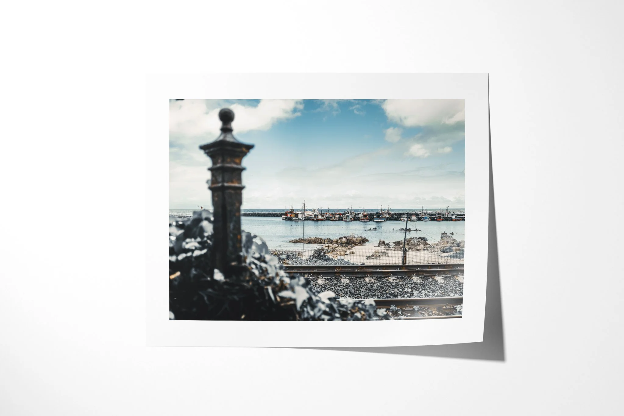 Unframed fine art paper print of Harbour, showing fishing boats moored across a calm harbour beyond a rocky shore and railway tracks, with a blurred iron post in front.