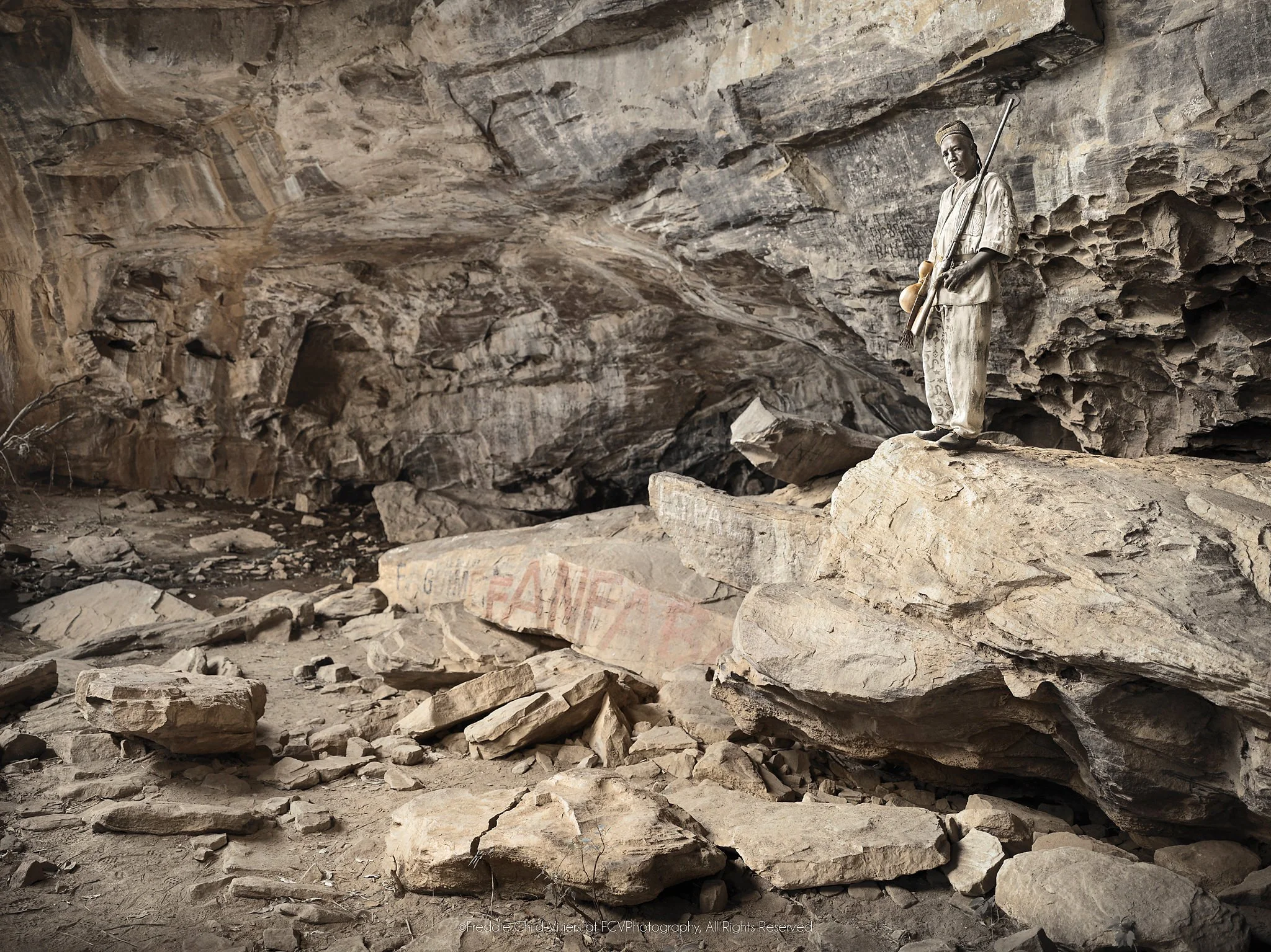 Bomary standing on a rock ledge within the broad chamber of a sacred cave.