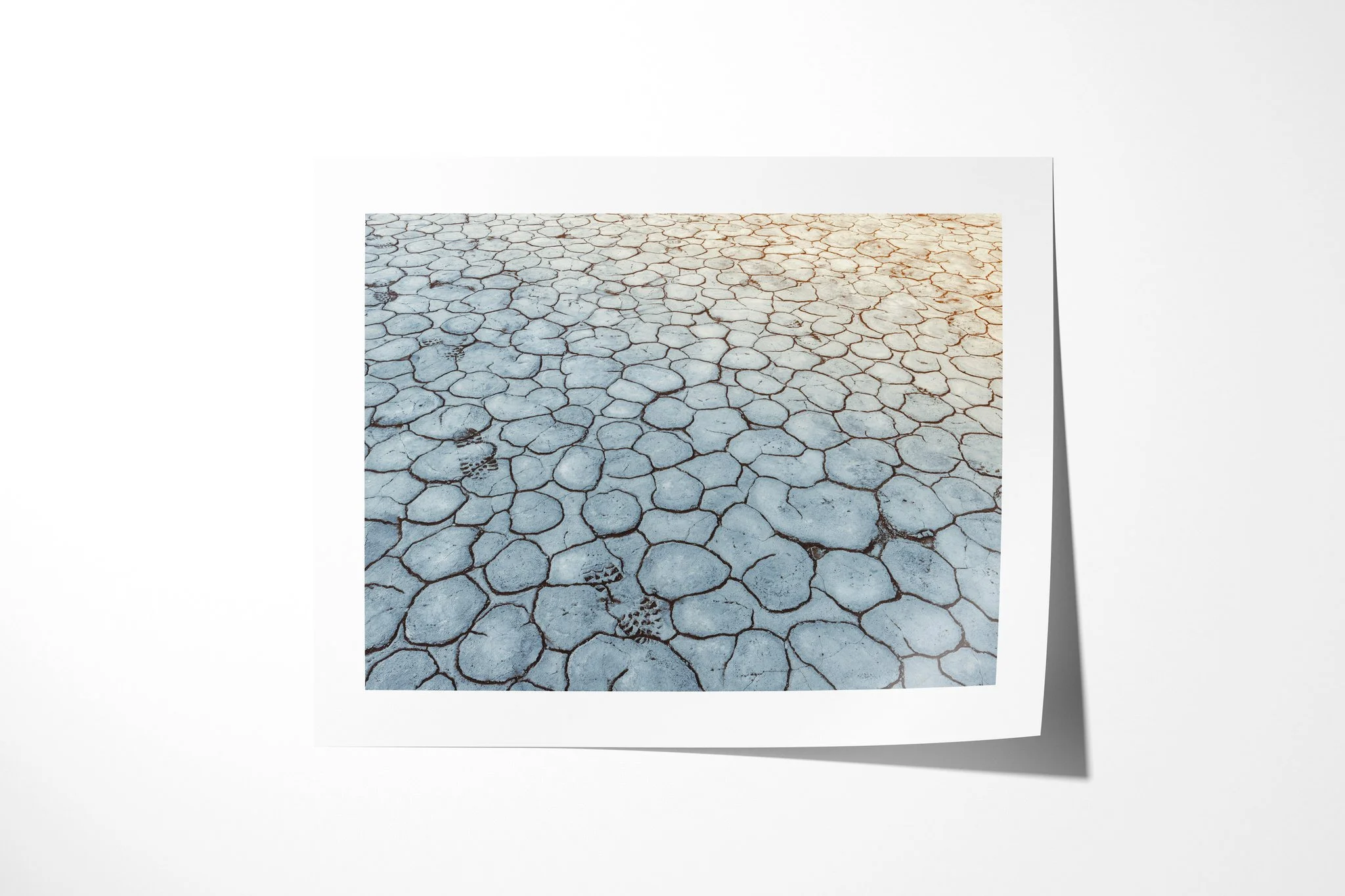 Footsteps presented as an unframed fine art paper print, depicting a trail of shoeprints crossing pale blue-grey cracked earth, with warm light entering from the upper right.