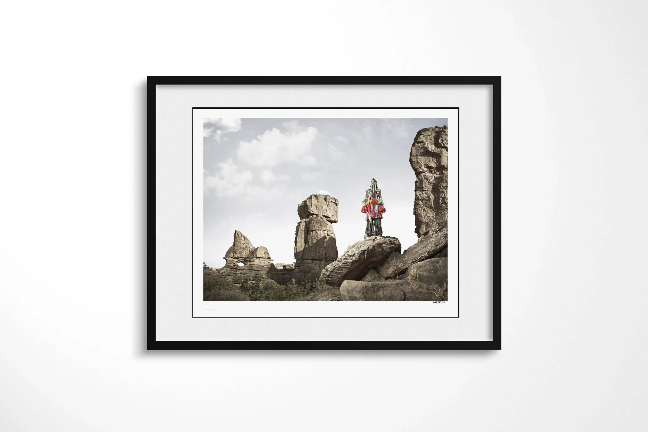 Framed presentation of Lagassa Lyai (Mask) in a black frame and white mount, showing a Dogon masked figure standing high among monumental sandstone formations.