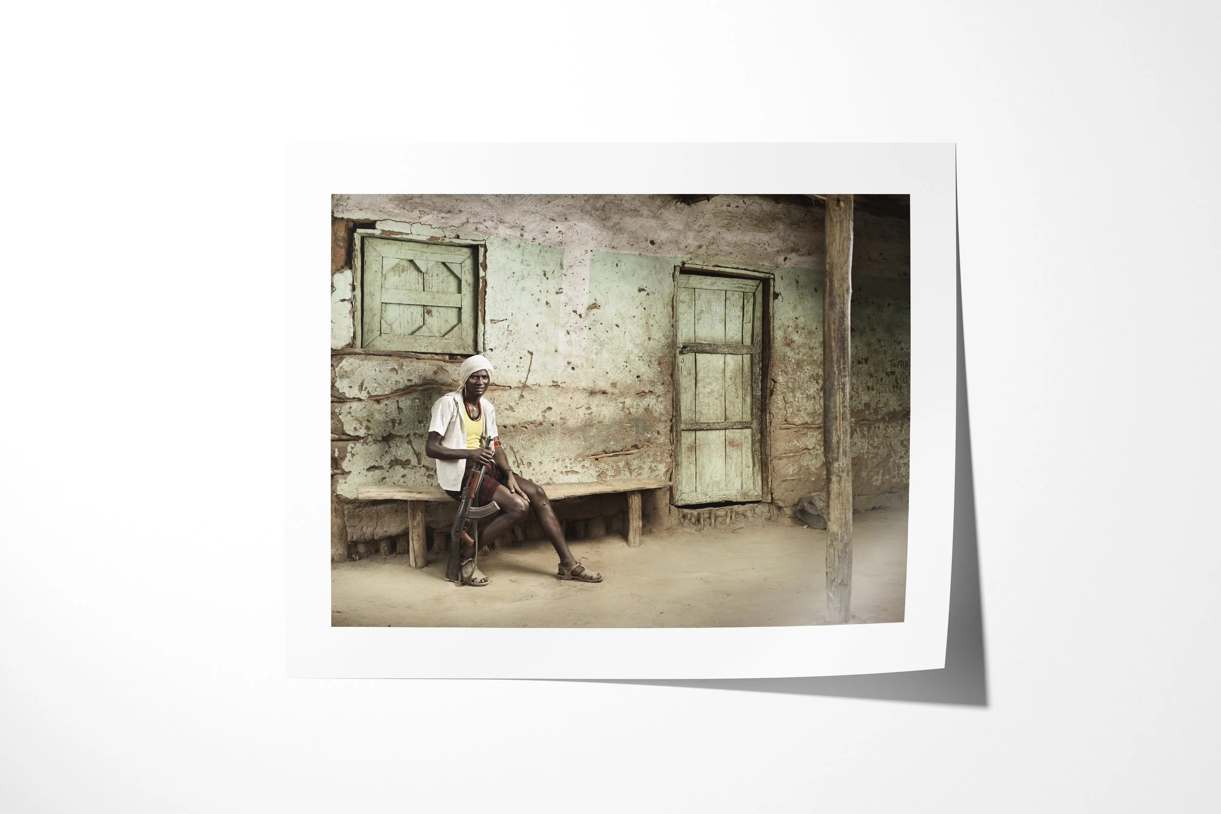 Fine art paper print of Arshal, showing an Arbore man seated with a rifle outside a weathered house with green shutters.