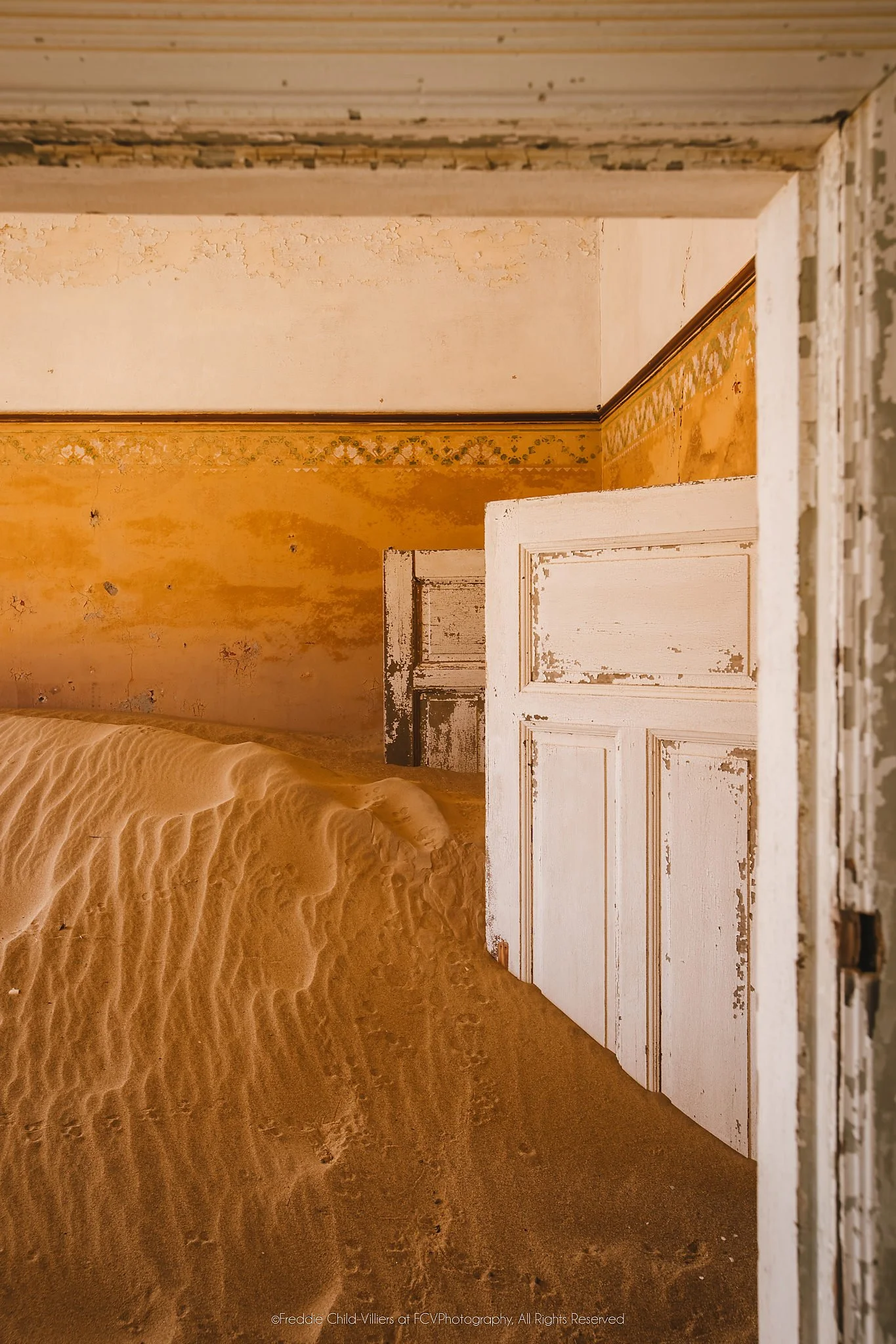 Chipped white doors and ochre walls layered behind a high drift of rippled sand in an abandoned interior.