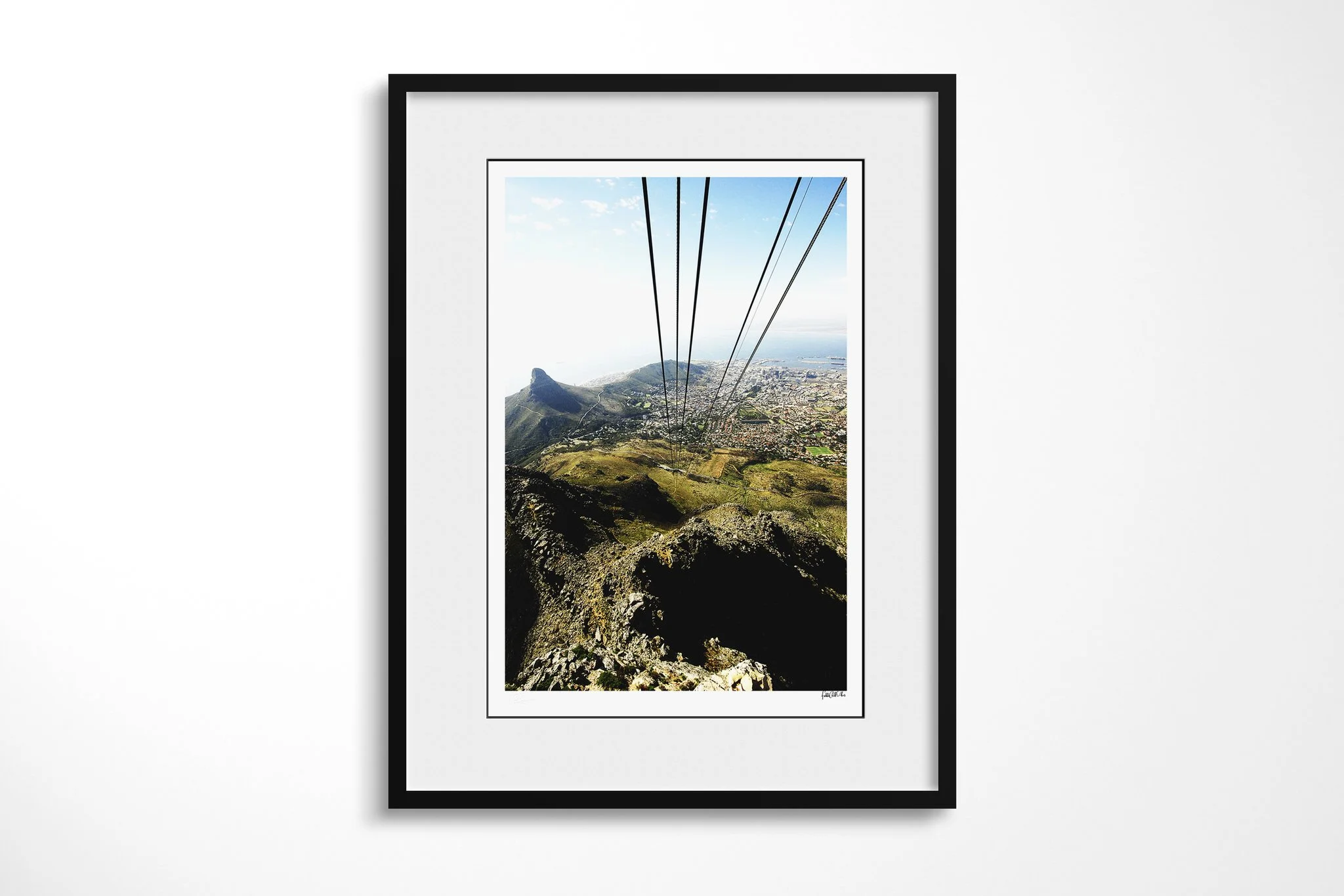 Framed presentation of Cape Town in a black frame and white mount, showing cable lines descending above Lion's Head, Cape Town's city bowl, harbour and Atlantic coast.