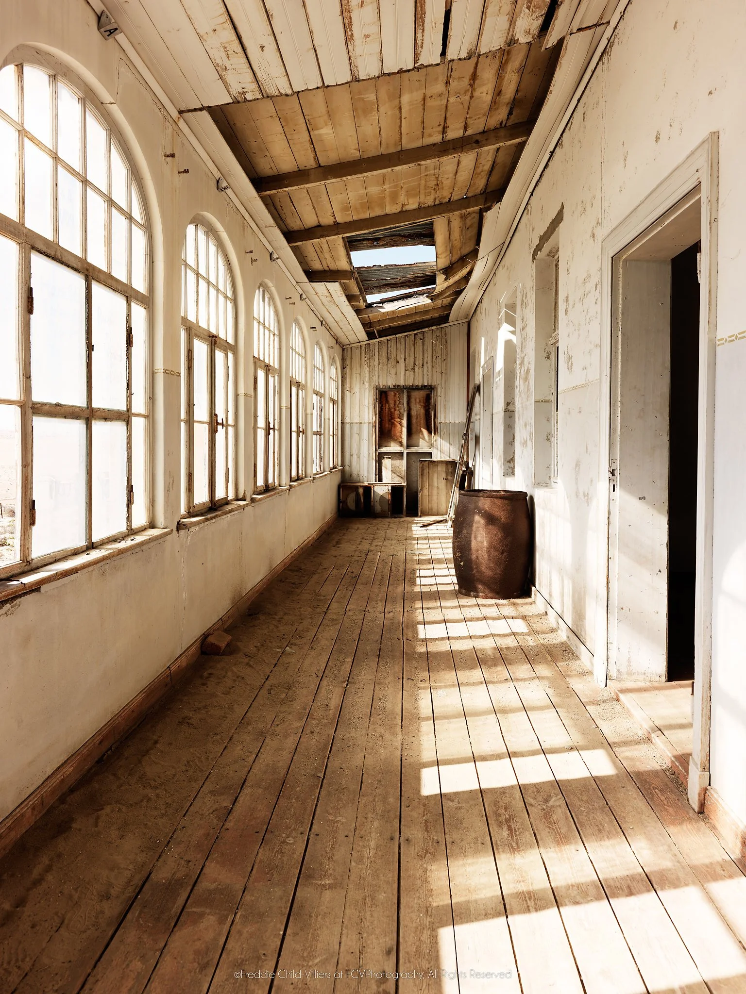 Long abandoned school corridor with arched windows, worn wooden floorboards, open doors and sunlight crossing peeling walls.