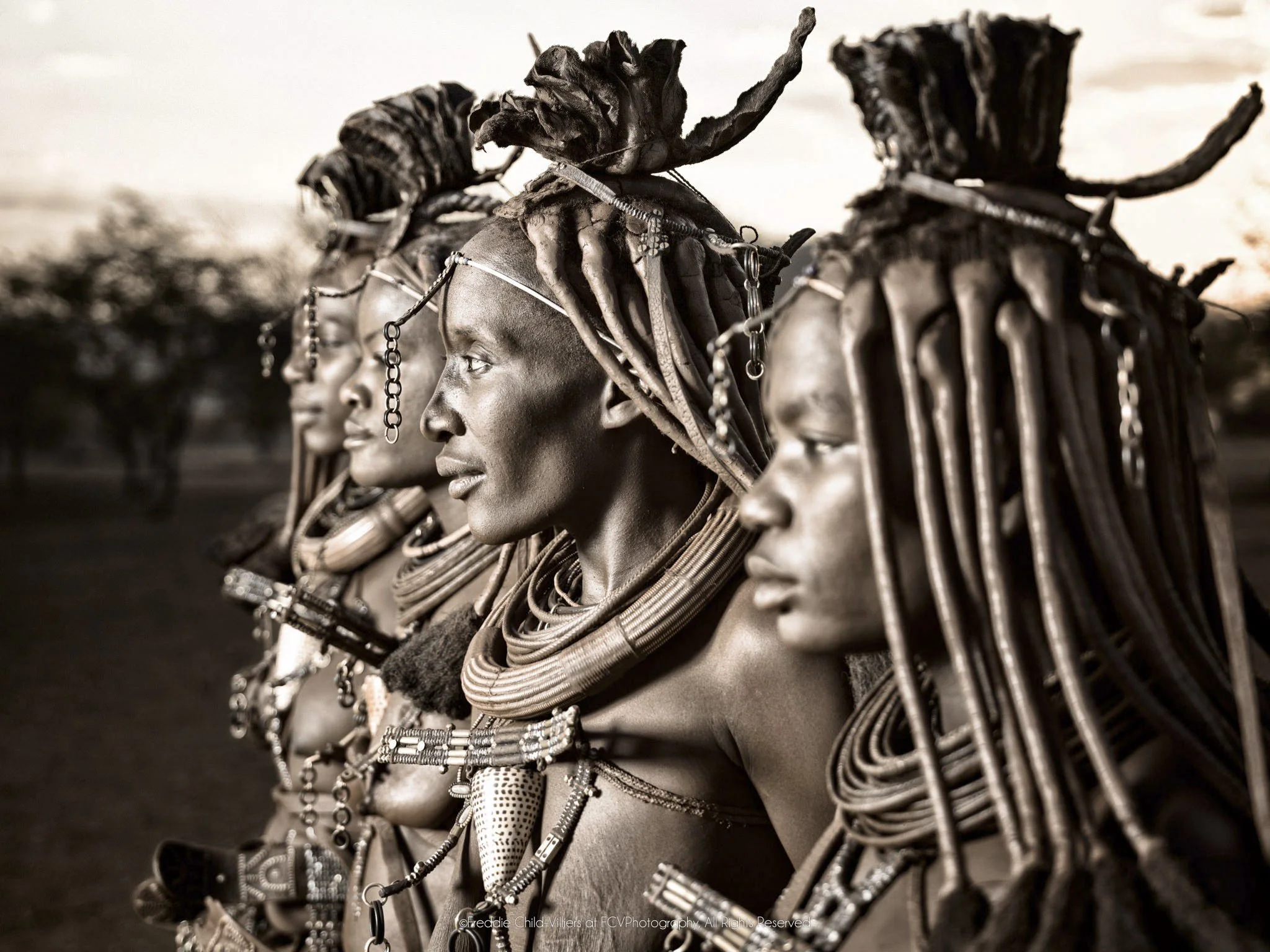 Mburaivaka (furthest), Waruakawa (far), Katitjii (focus) and Kayeripewa (closest), four Himba women shown in close profile, their otjize-dressed hair and jewellery overlapping across the frame.