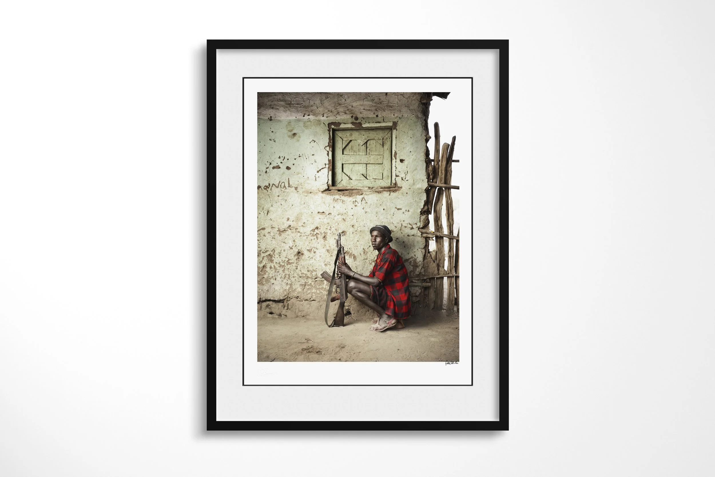 Framed presentation of Hegere in a black frame and white mount, showing an Arbore man seated on a low wooden headrest-stool with a rifle beside an earthen wall.