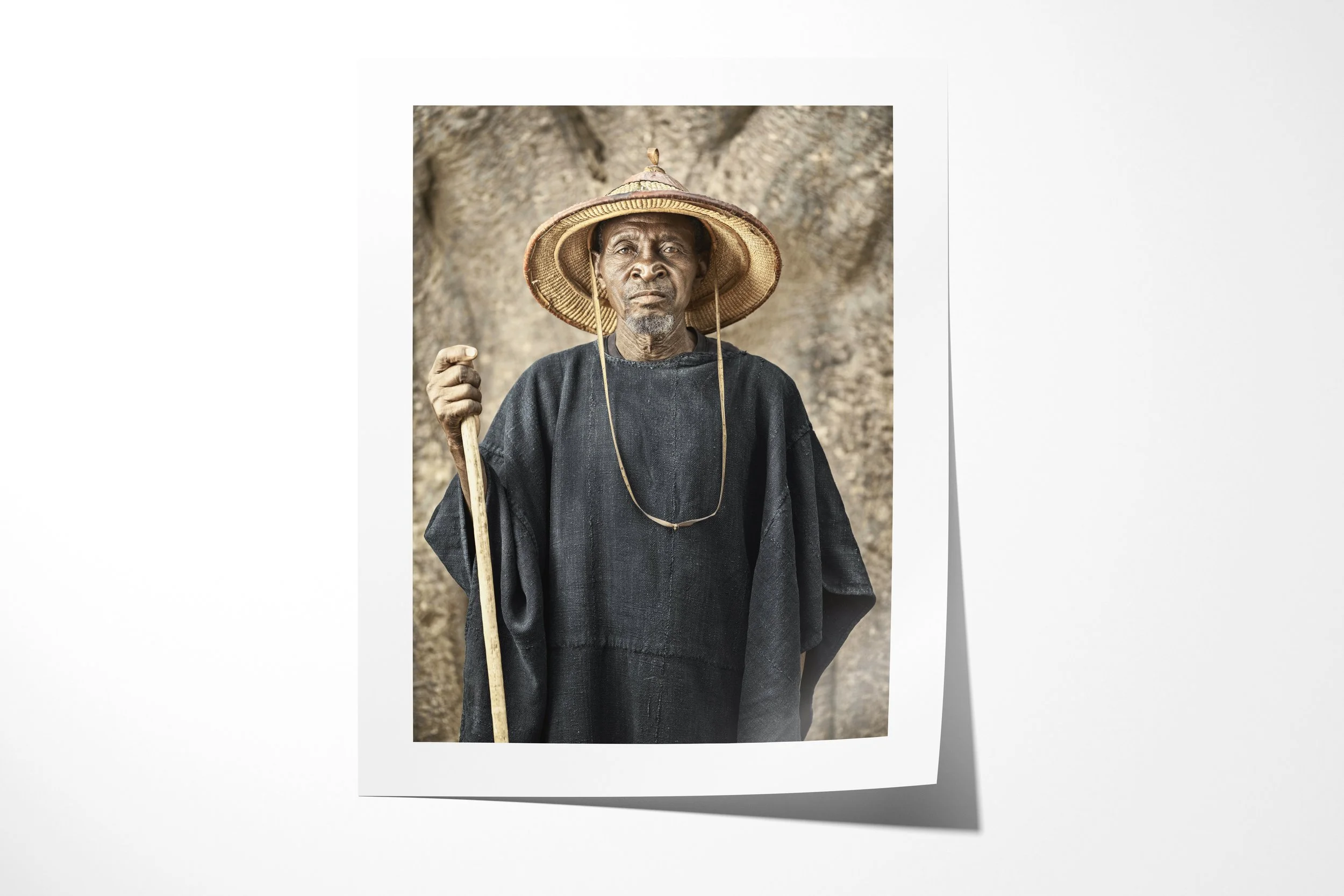 Fine art paper print of Tigué, showing Tigué in a dark robe and broad straw hat holding a staff before a baobab trunk.