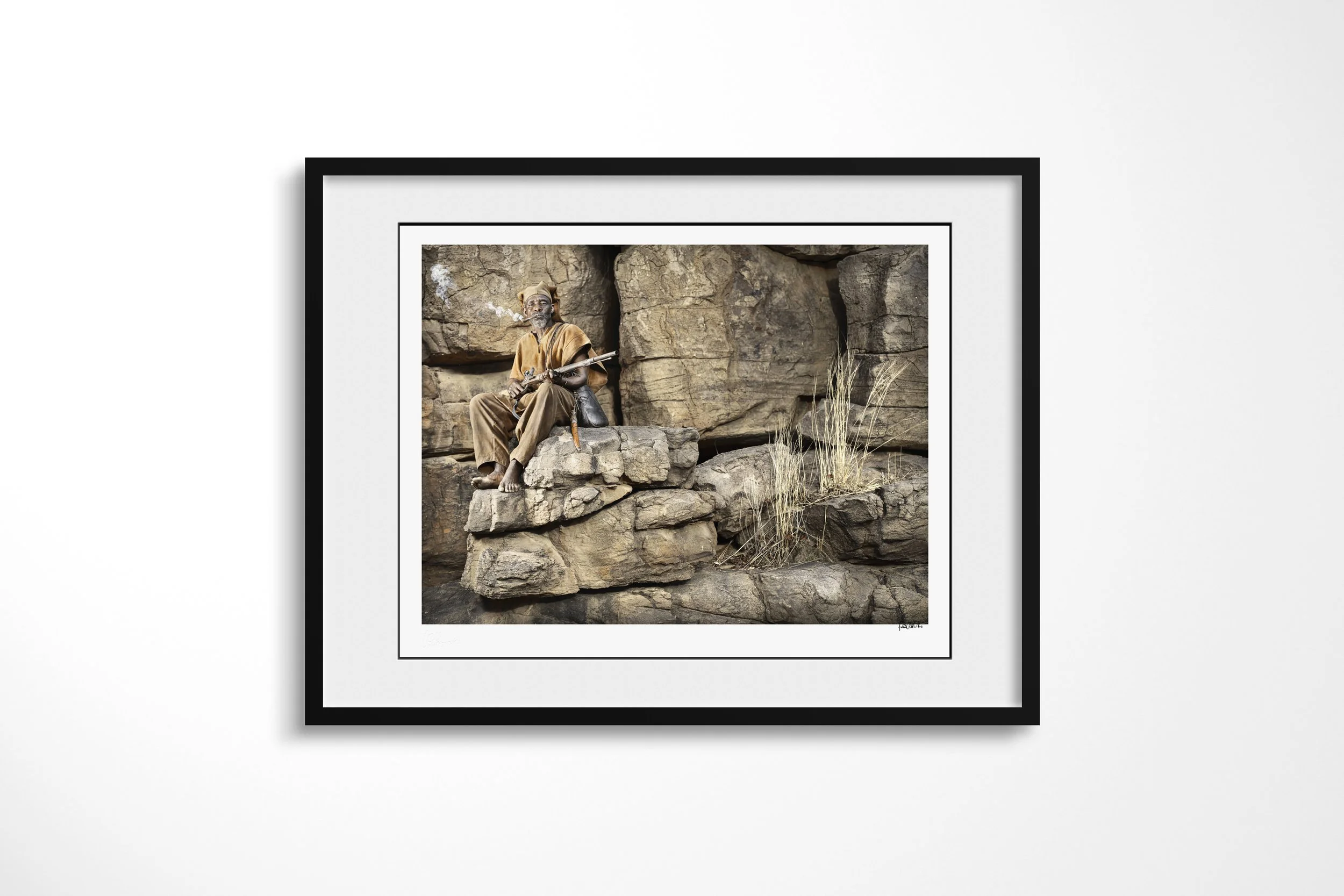 Framed presentation of Antandou in a black frame and white mount, showing Antandou seated on layered rock with a rifle as pipe smoke rises beside him.