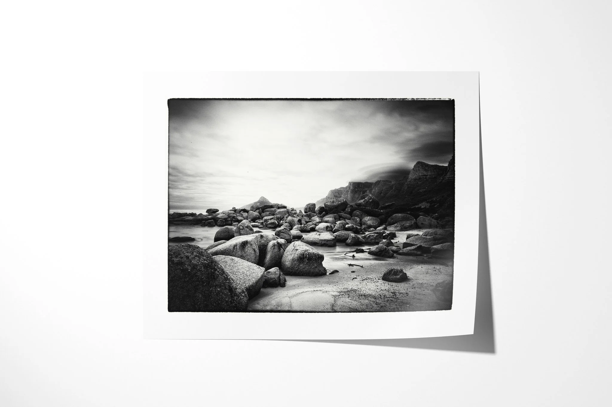 Twelve Apostles presented as an unframed fine art paper print, depicting a black-and-white boulder beach beneath Cape Town's Twelve Apostles cliffs, with low cloud and softened seawater.