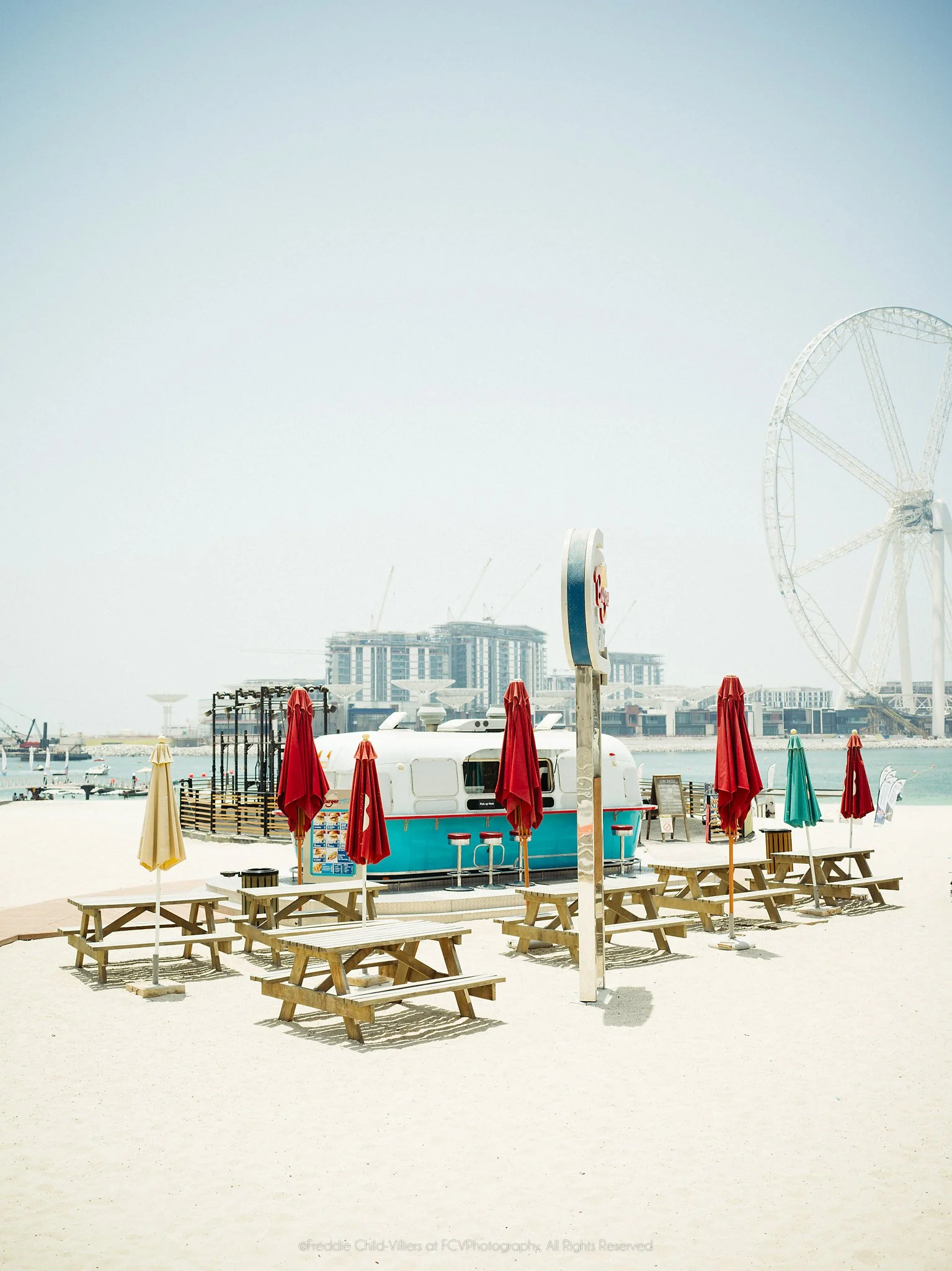 Empty picnic tables and closed colourful parasols beside a small waterfront kiosk, with an observation wheel in the distance.
