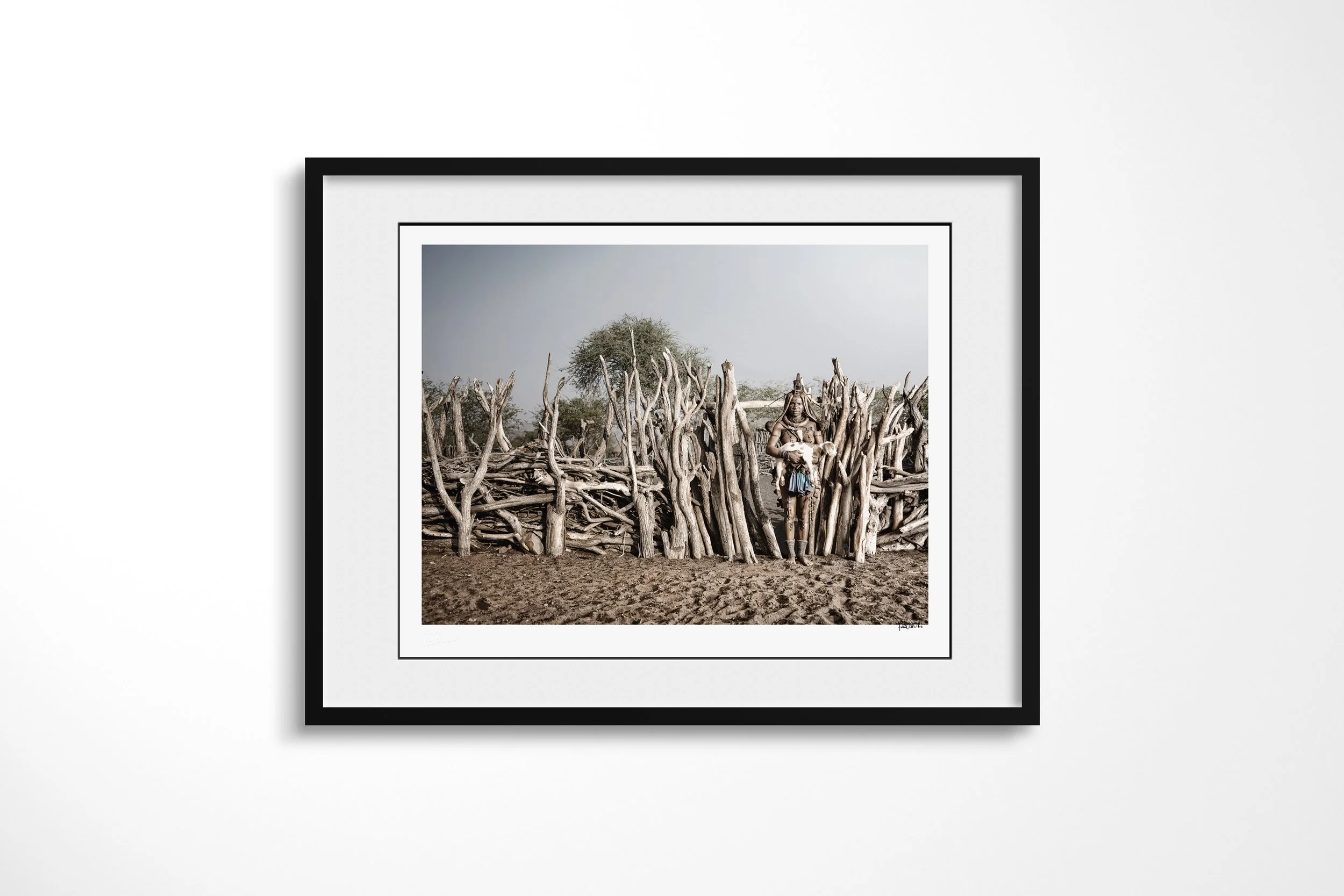 Framed presentation of Twahenwa III in a black frame and white mount, showing Twahenwa holding a young goat beside the branch enclosure of a Himba homestead.