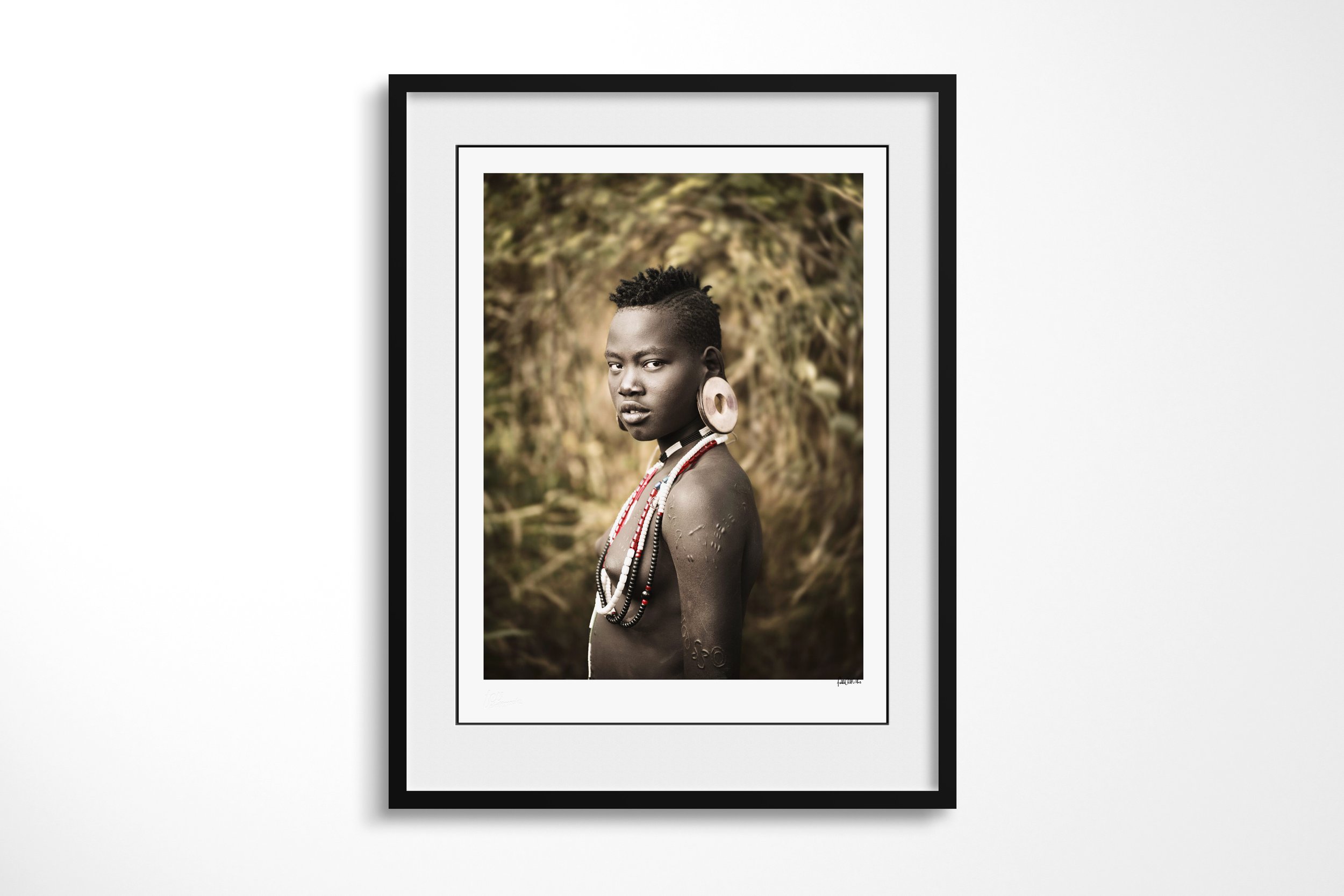 Framed presentation of Kernahoir in a black frame and white mount, showing a young Mursi girl in profile with a large ear disc, bead necklaces and scarification on her shoulder.
