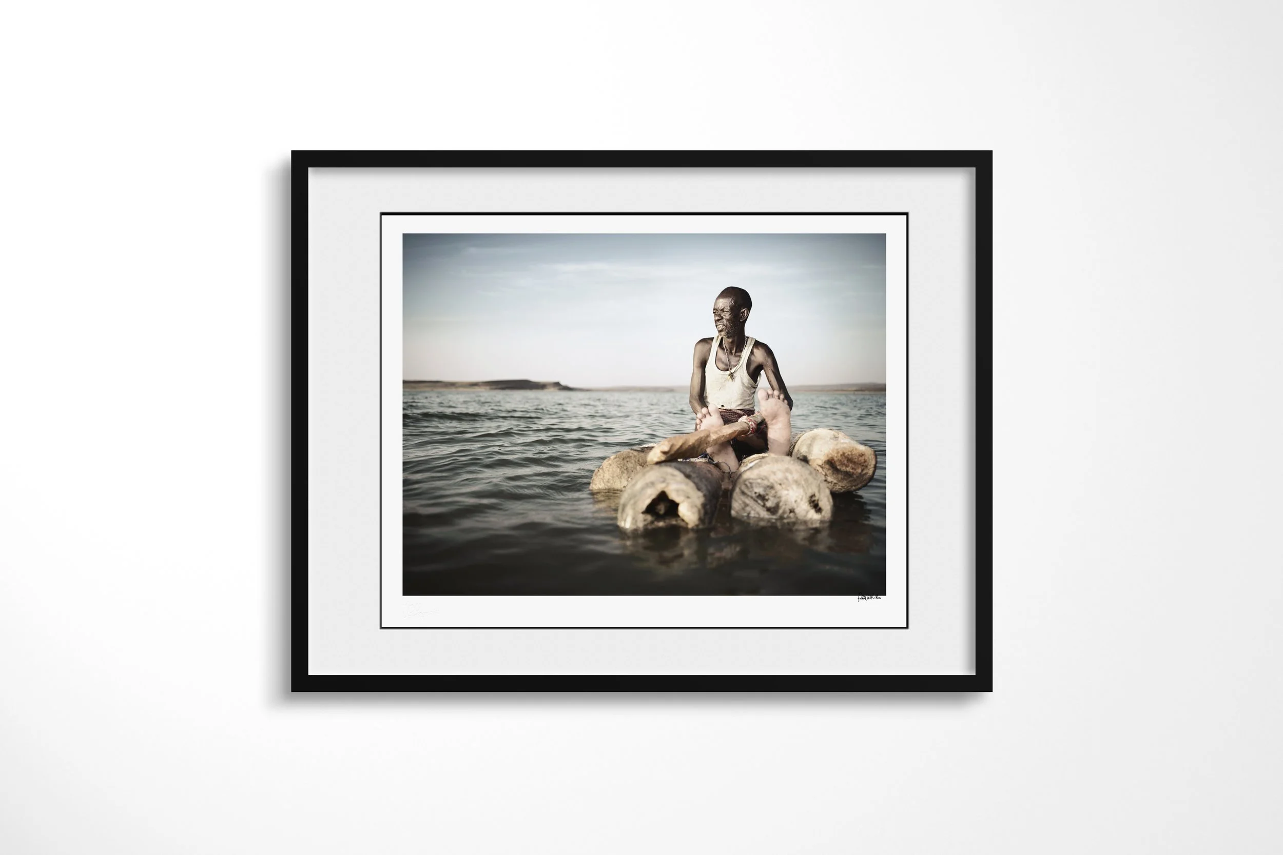 Framed presentation of Lengutuk II in a black frame and white mount, showing an El Molo fisherman seated on a raft of lashed palm logs on Lake Turkana.