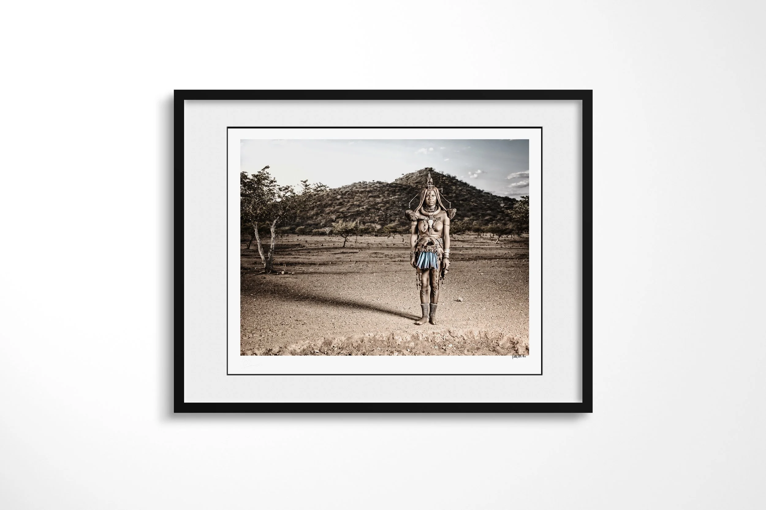 Framed presentation of Twahenwa in a black frame and white mount, showing Twahenwa in elaborate ceremonial dress standing in open country before low hills.