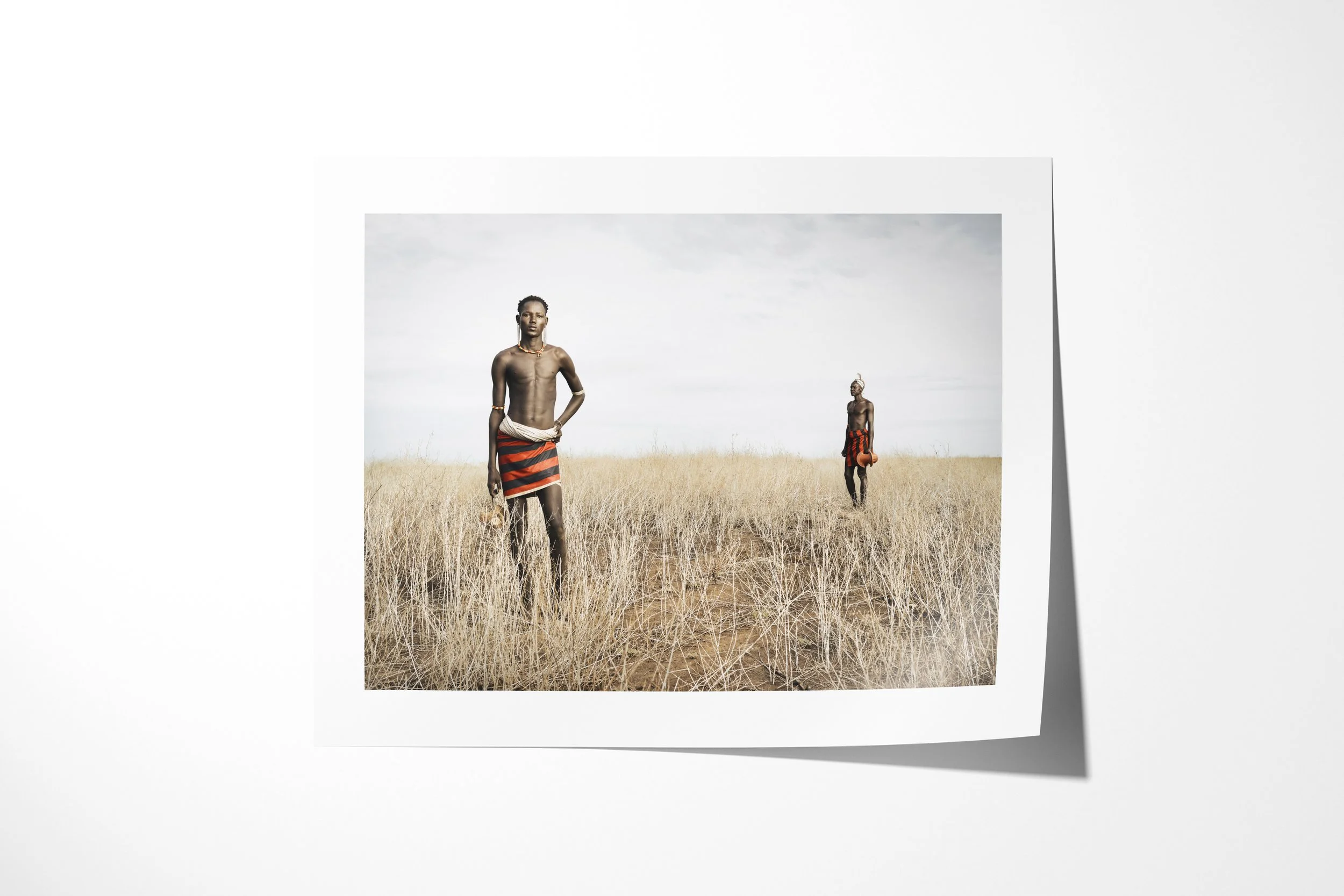 Fine art paper print of Lokoloi & Losiakou, showing two Daasanach men in red-and-black wraps standing apart in tall dry grass.