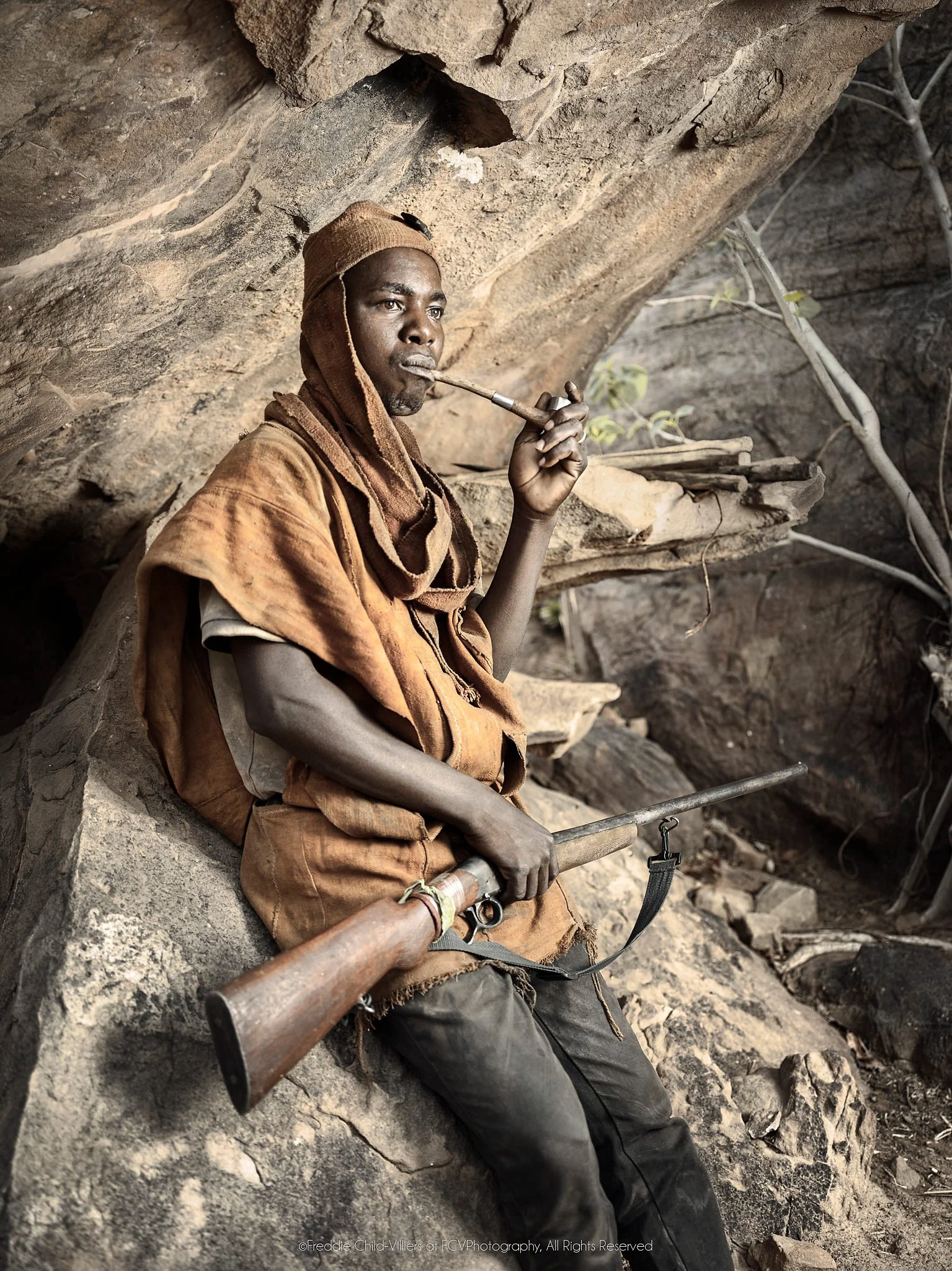 Anbile seated in a rocky shelter smoking a long pipe with a rifle across his lap.