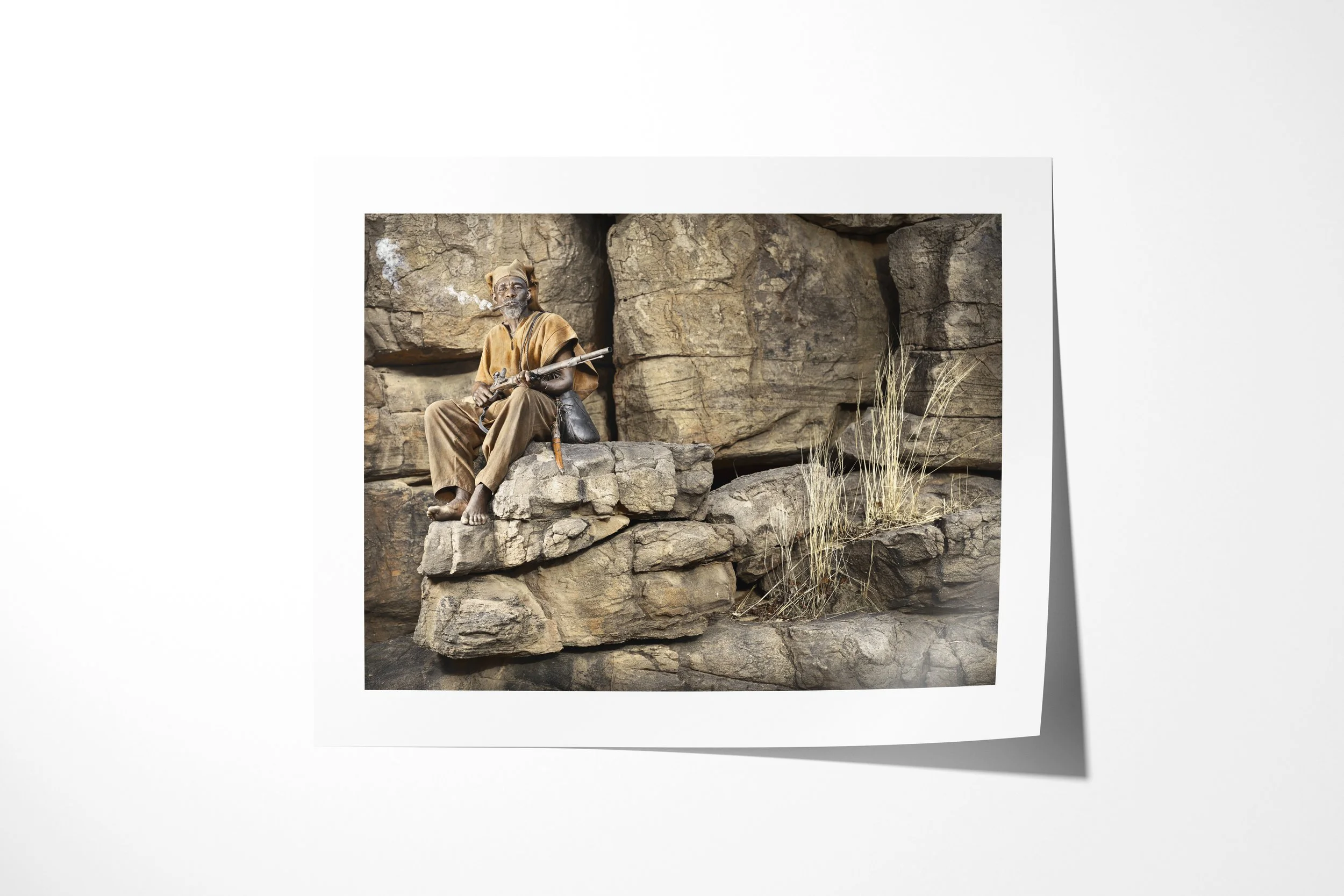 Fine art paper print of Antandou, showing Antandou seated on layered rock with a rifle as pipe smoke rises beside him.