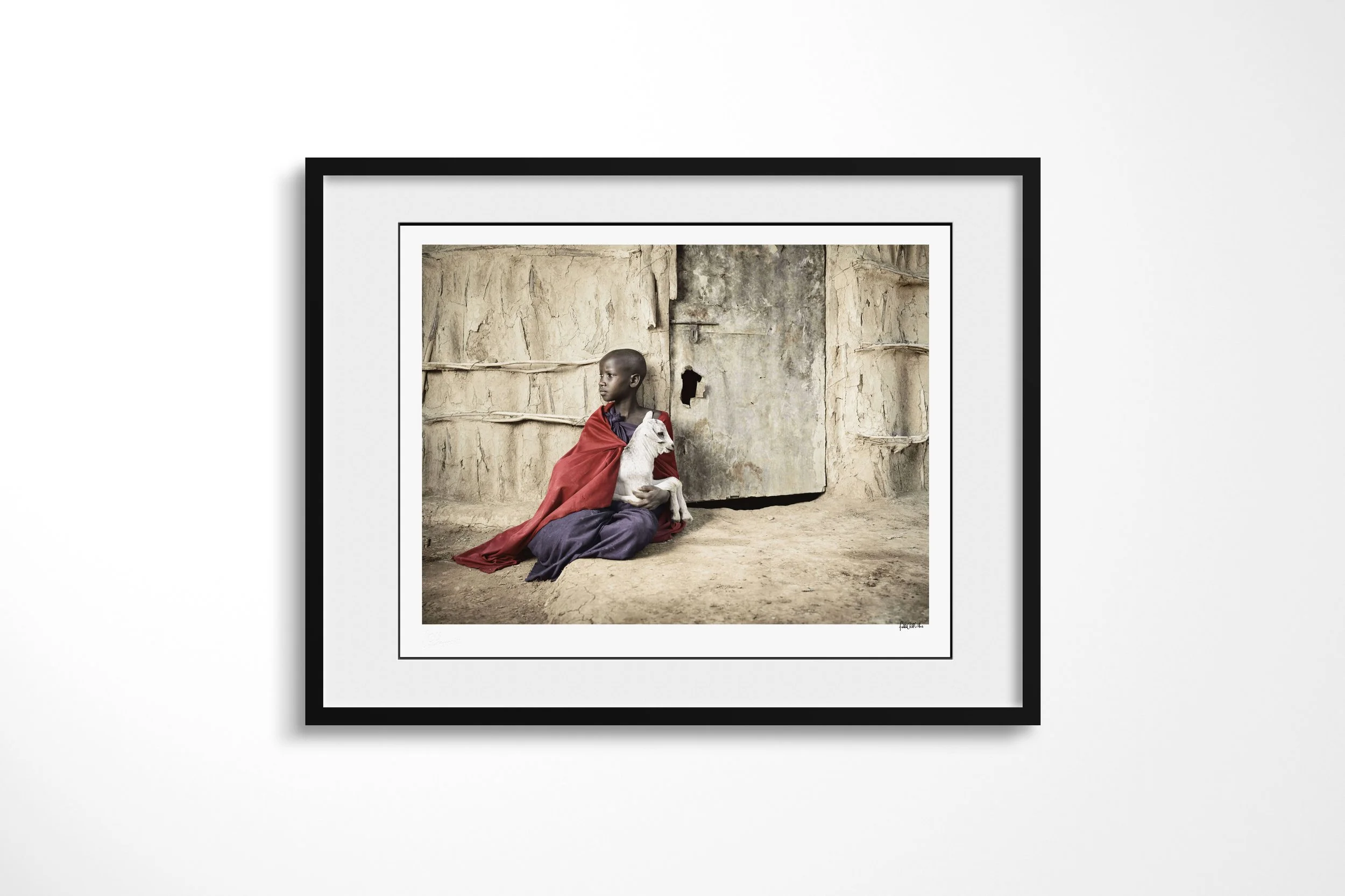 Framed presentation of Nawasa in a black frame and white mount, showing a Maasai girl wrapped in red cloth holding a young white goat outside an earthen house.
