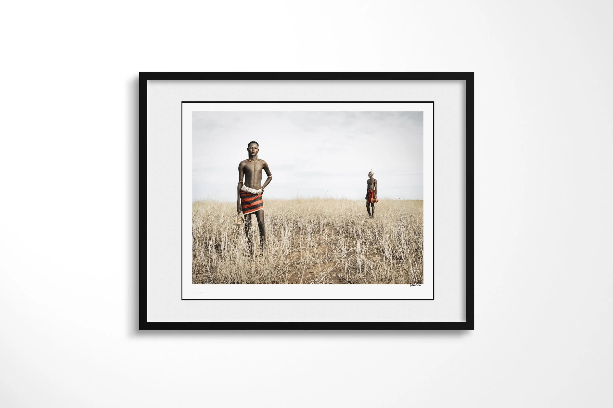 Framed presentation of Lokoloi & Losiakou in a black frame and white mount, showing two Daasanach men in red-and-black wraps standing apart in tall dry grass.