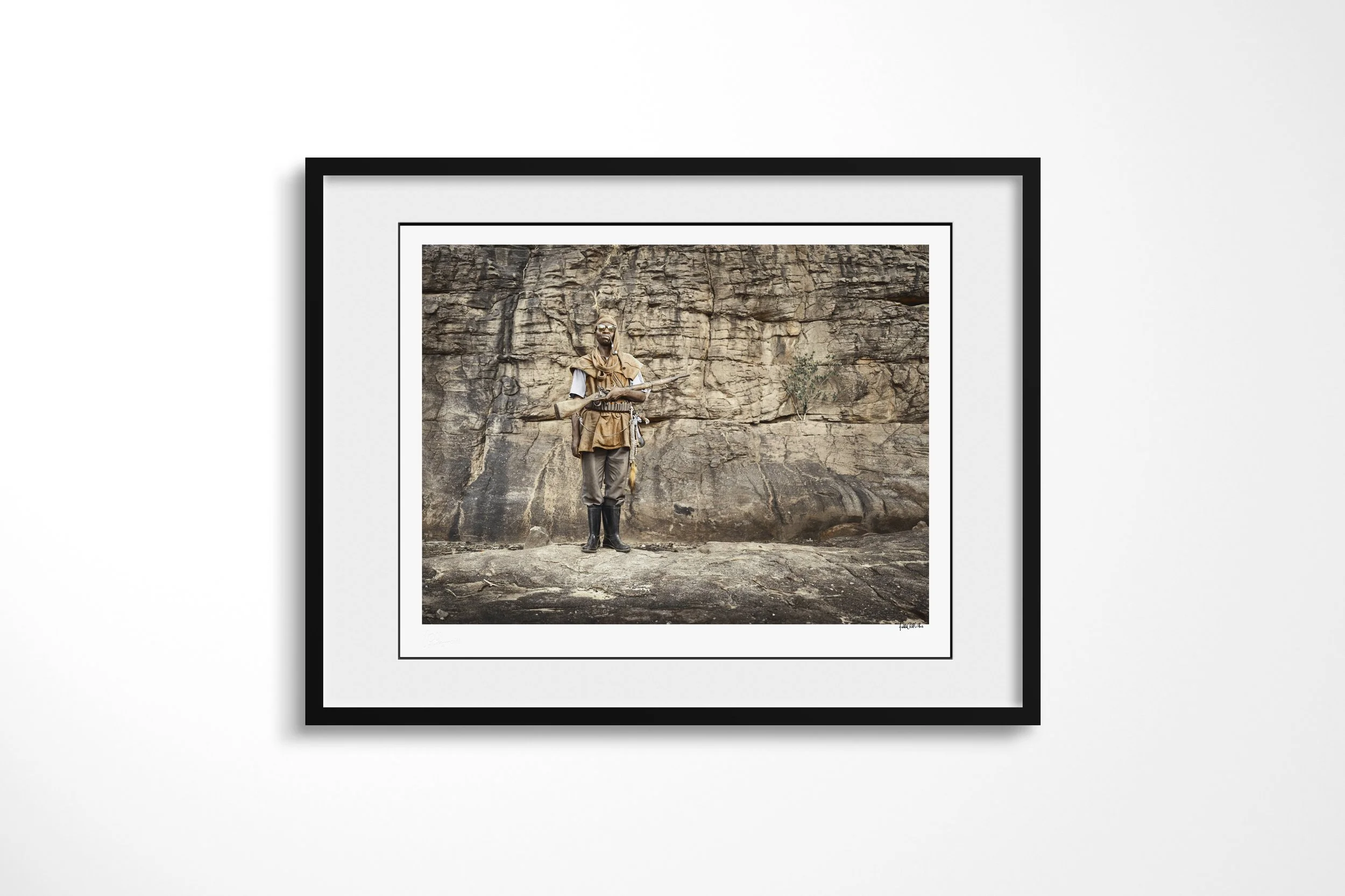 Framed presentation of Diongo in a black frame and white mount, showing Diongo standing with a rifle, ammunition belt and boots before a rock wall.