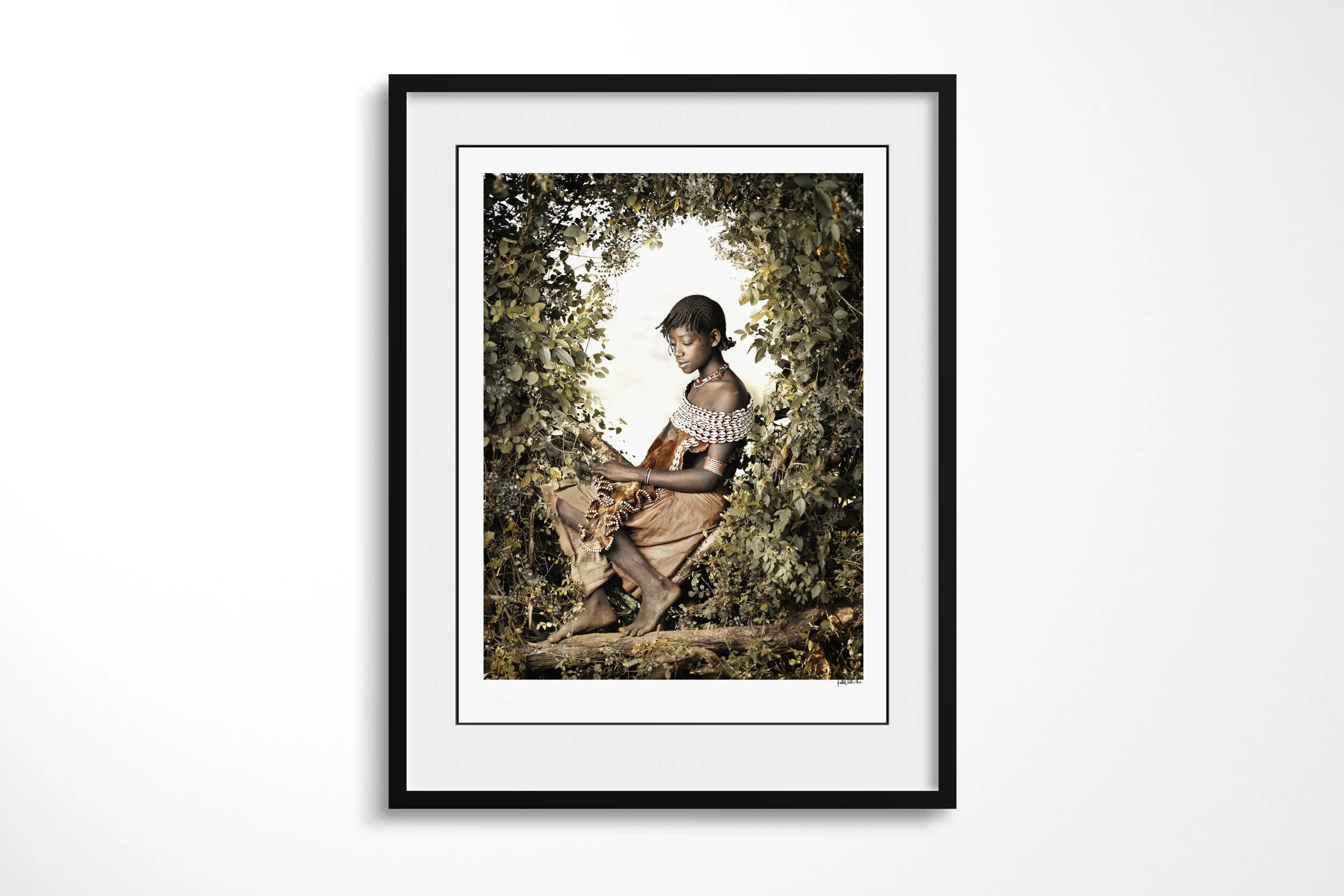 Framed presentation of Semba in a black frame and white mount, showing a young Banna woman seated in foliage wearing layered beadwork and a cowrie-covered leather garment.