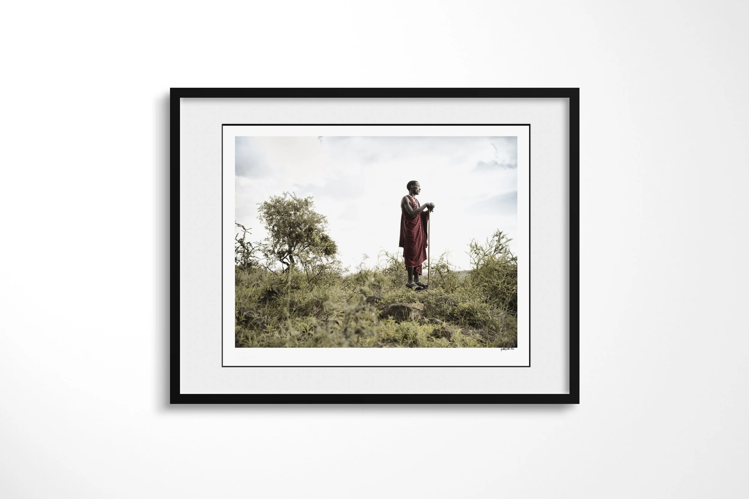 Framed presentation of Loboe in a black frame and white mount, showing a Maasai man with a staff standing on a low rise amid dense green scrub.