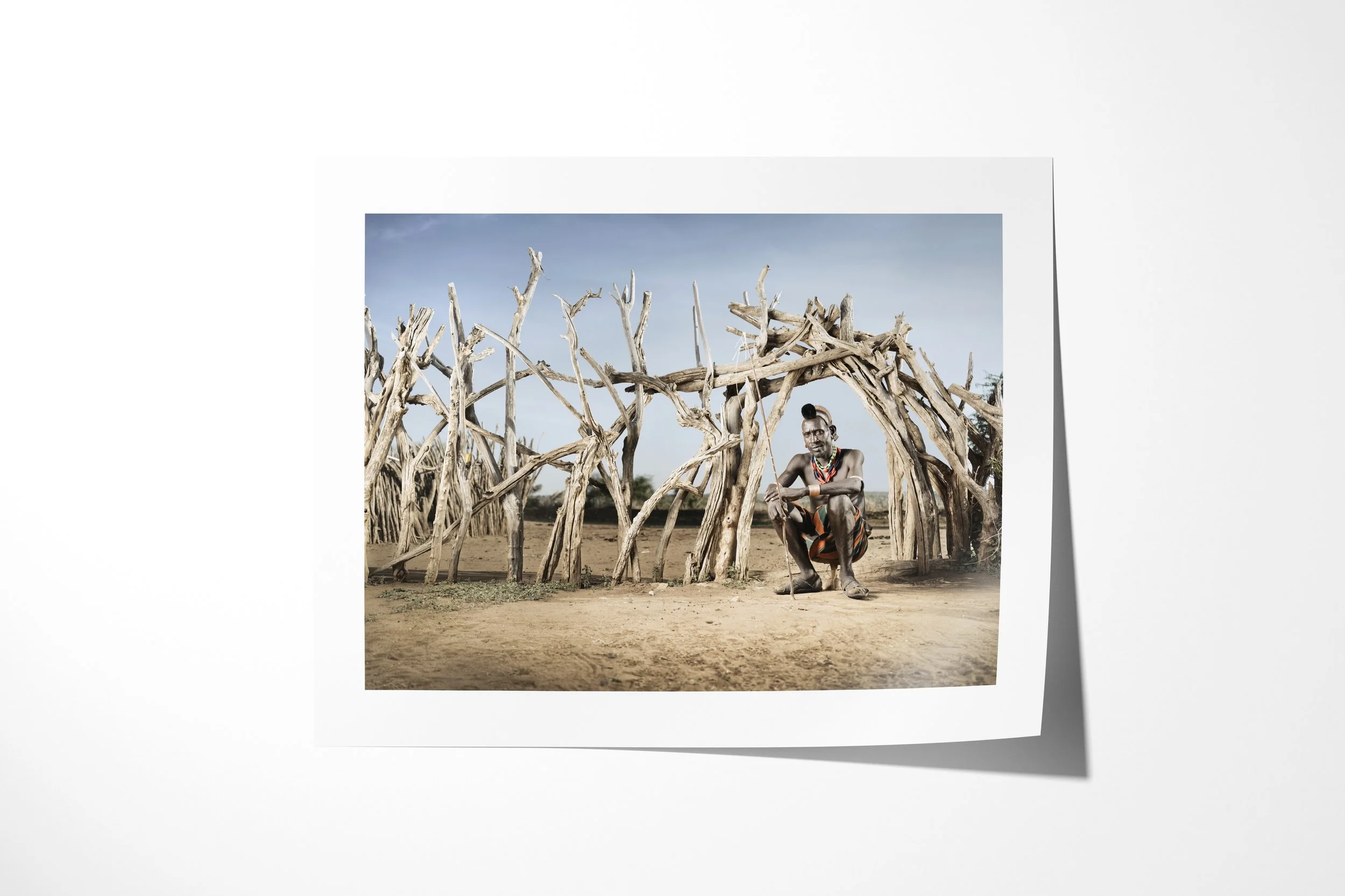 Fine art paper print of Woro, showing an older Hamar man with pale body paint and layered beadwork crouching inside a branch-built enclosure.