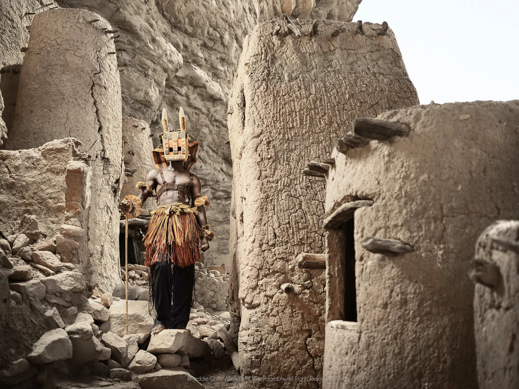 Djomo (Mask), a Dogon Djomo mask moving through a narrow passage between earthen and stone buildings.