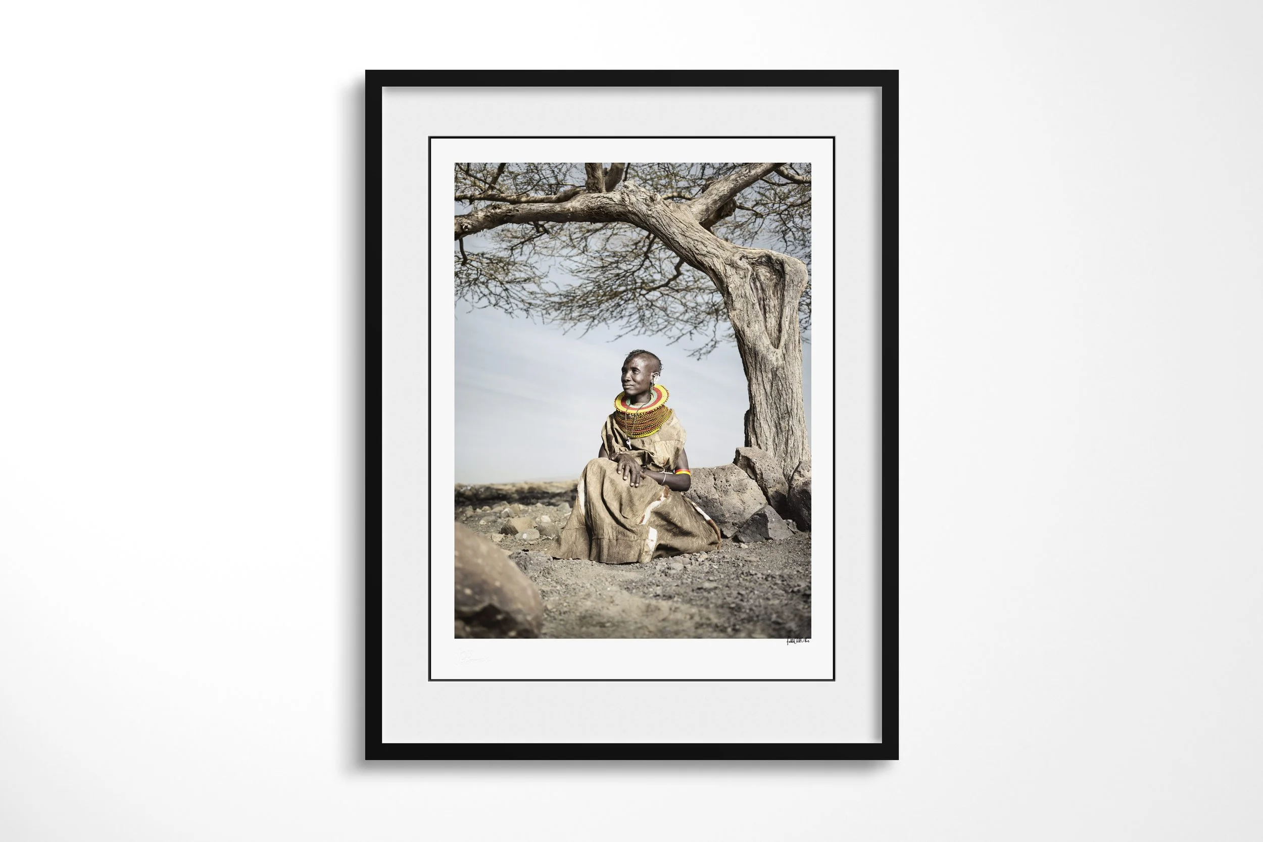 Framed presentation of Lotilan in a black frame and white mount, showing a Turkana woman in a hide dress seated beneath an acacia in rocky country.