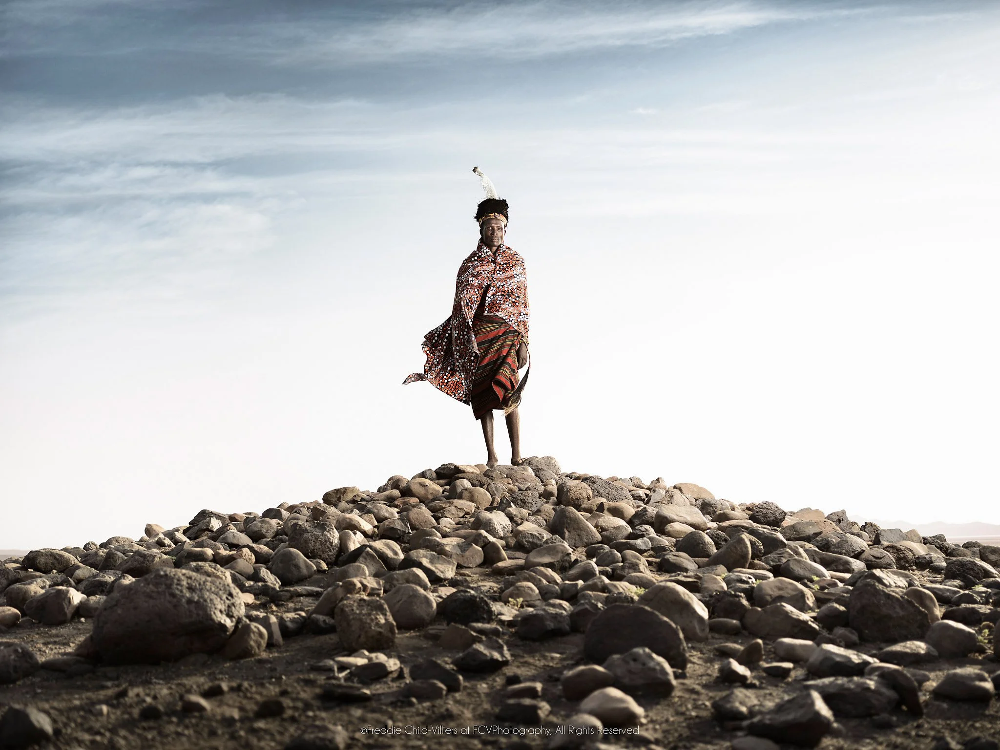 Lomus standing on a mound of rounded rocks as wind lifts his patterned cloth.