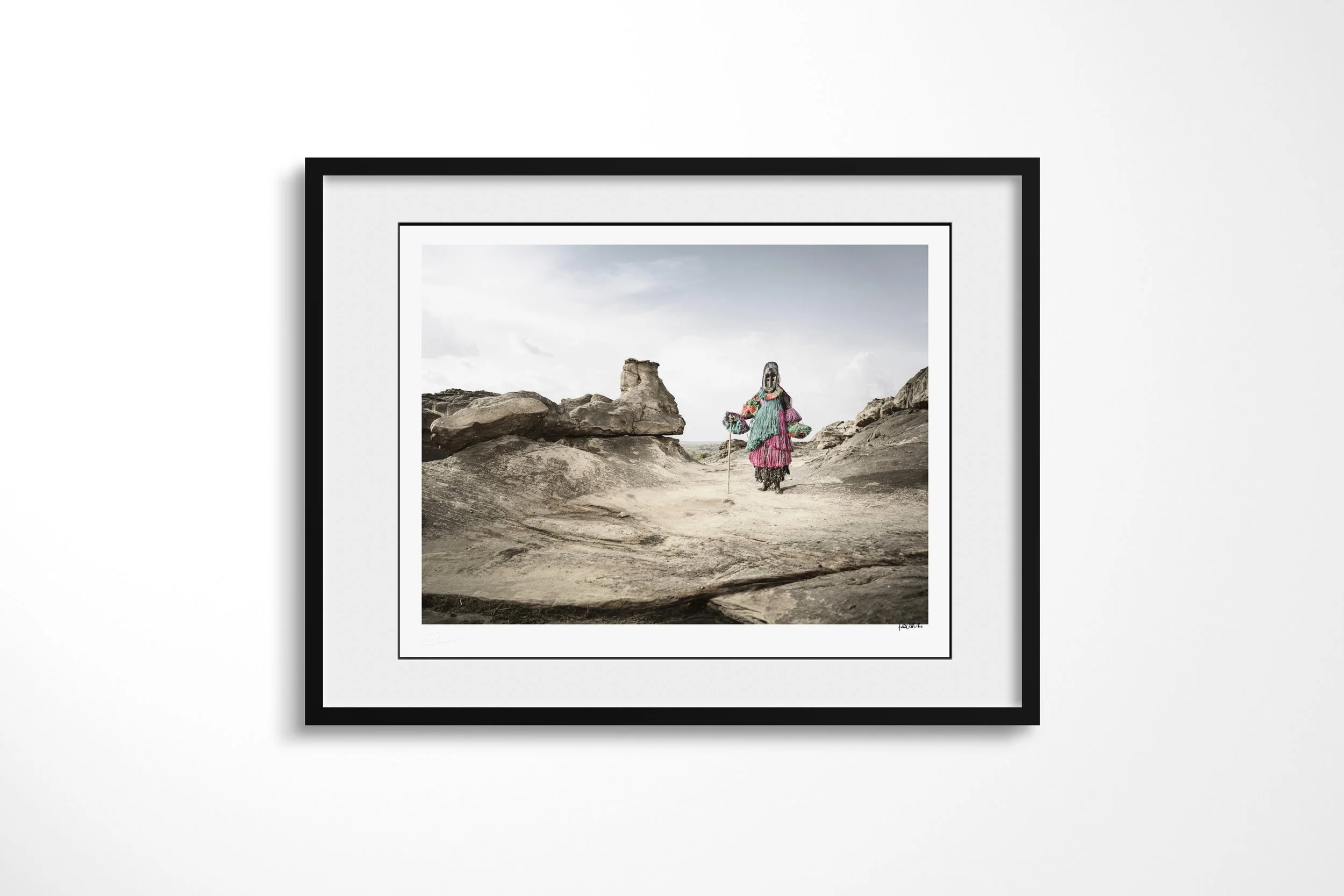 Framed presentation of Dana (Mask) in a black frame and white mount, showing a Dogon hunter mask walking across open rock beneath a pale sky.