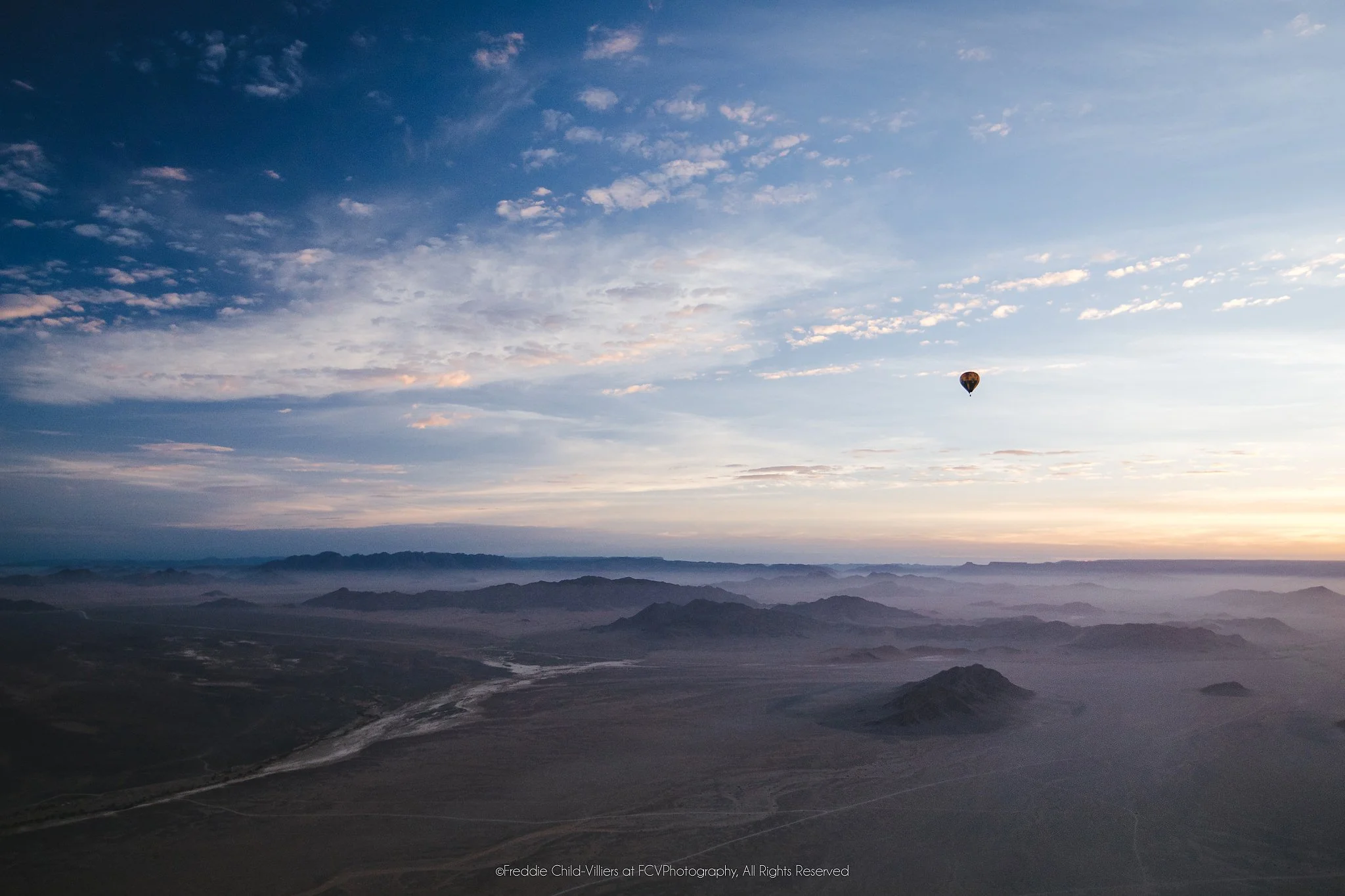 Small hot-air balloon above an expansive arid landscape of ridges, valleys and pale winding channels at dawn.