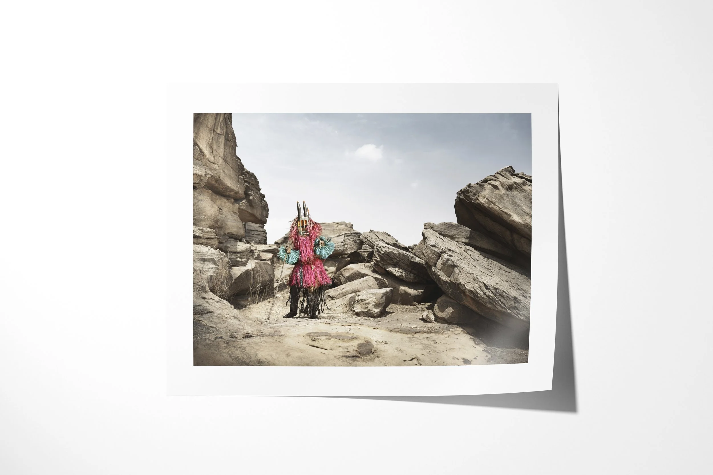 Fine art paper print of Walu (Mask), showing a Dogon masked figure in bright fibre costume standing in a narrow rocky passage.