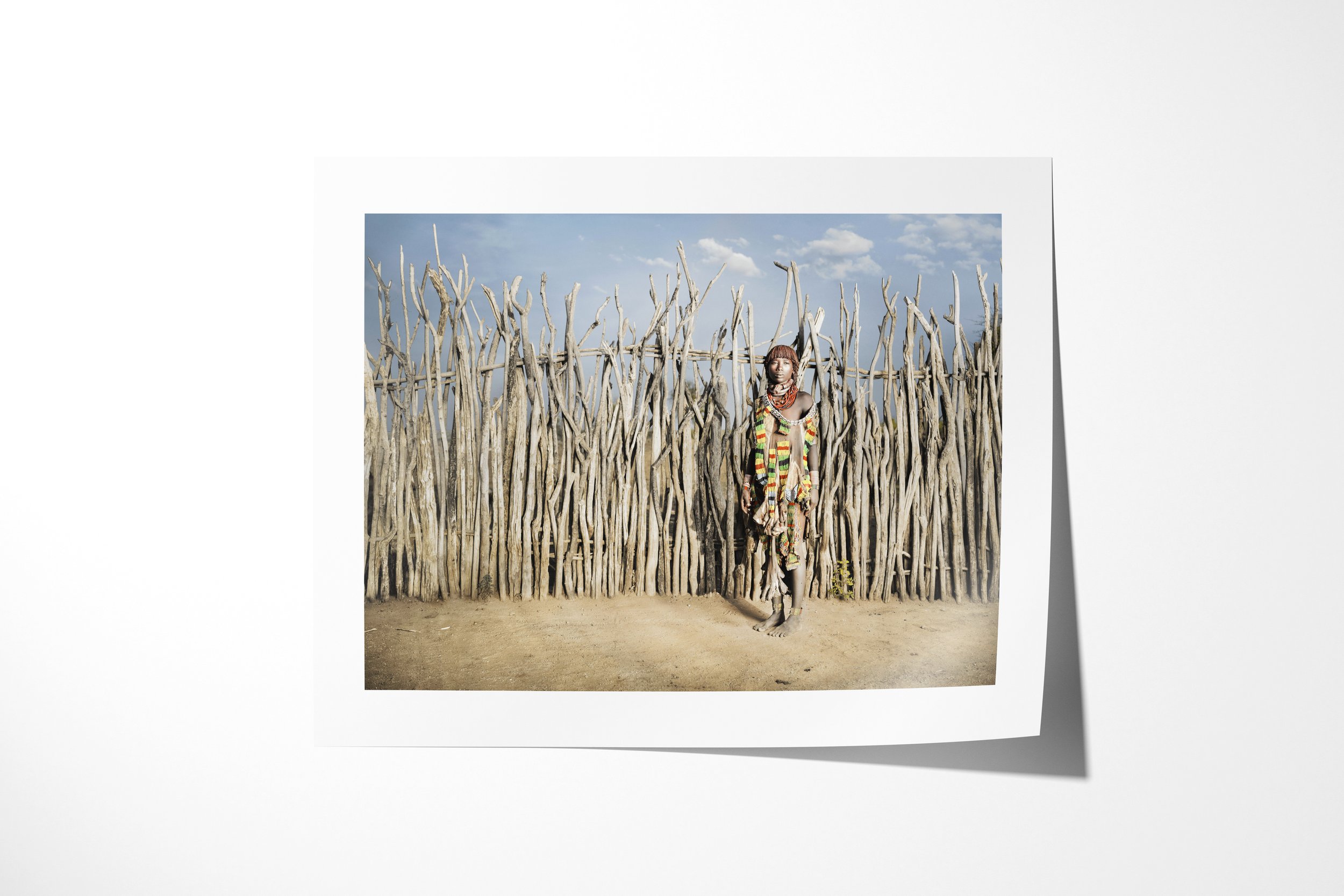 Fine art paper print of Aito, showing a Hamar woman in layered beadwork and decorated hide clothing standing against a tall wooden fence.