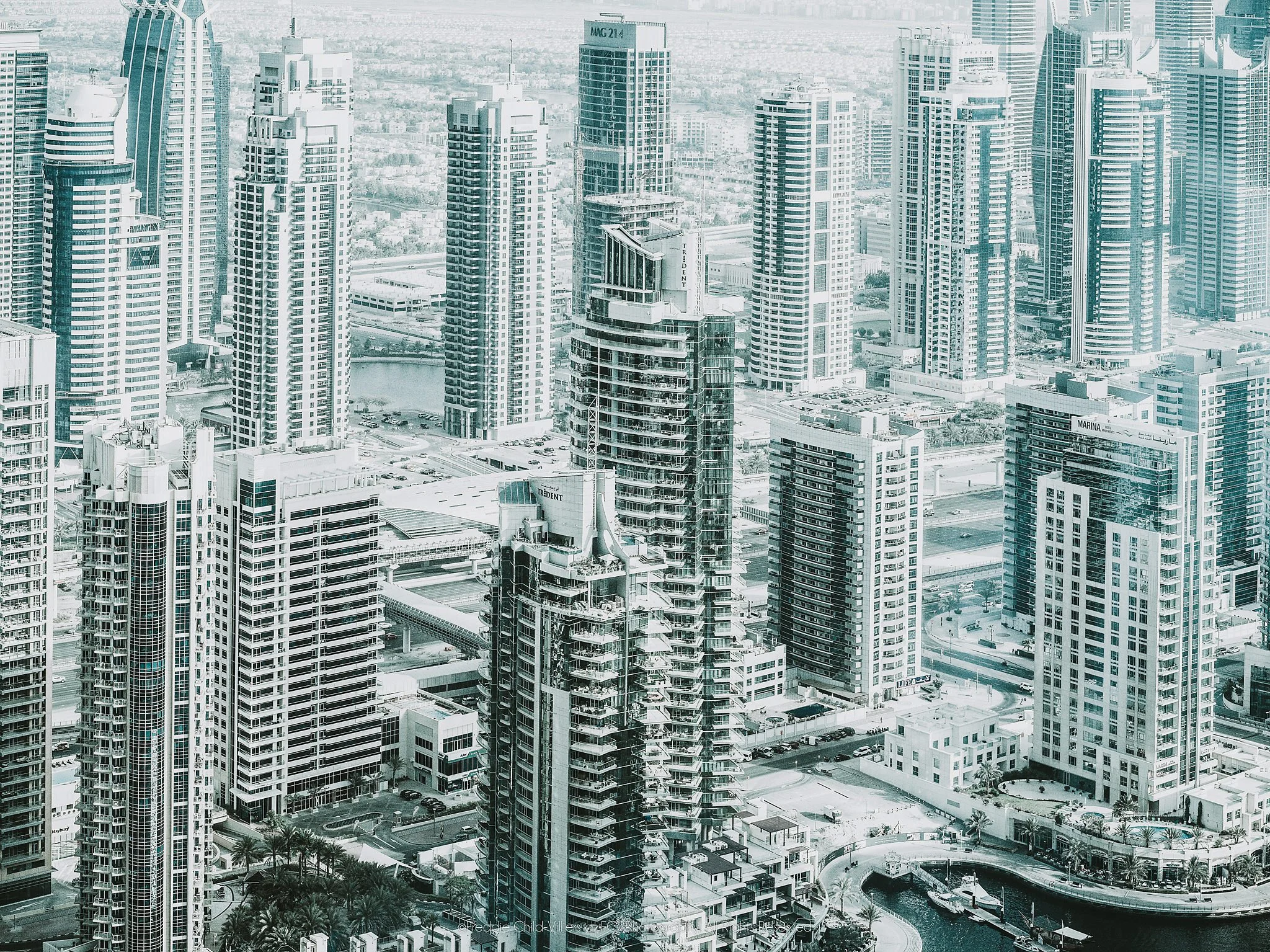 Dense modern cityscape of high-rise towers, balconies, roads and waterways seen from above in cool pale tones.