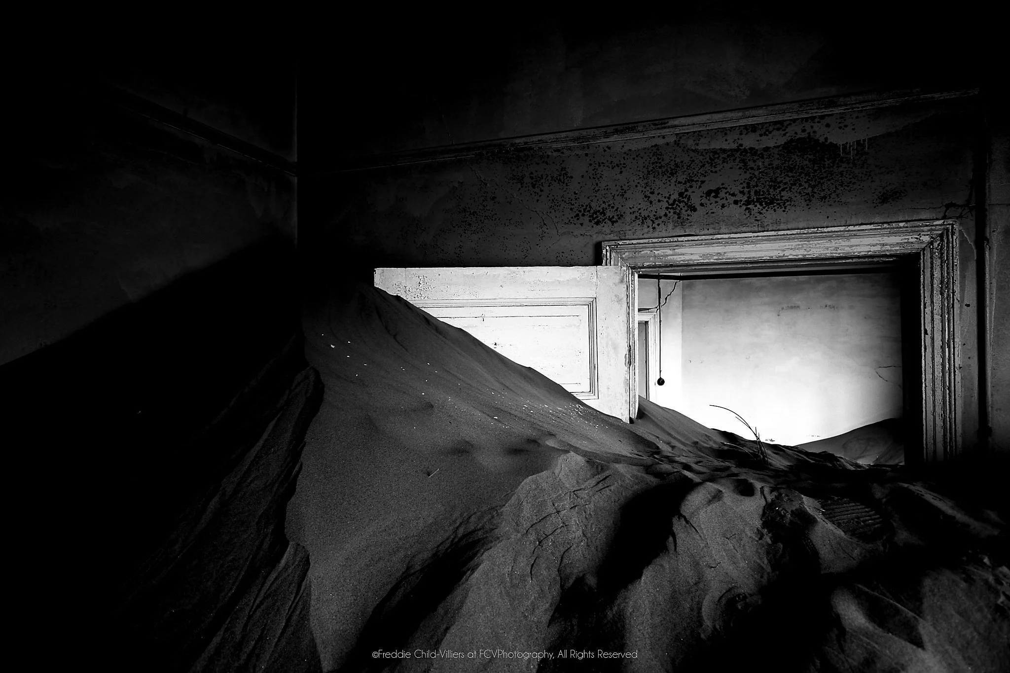 Steep sand drift filling a dark abandoned room and rising through a partly open doorway in black and white.