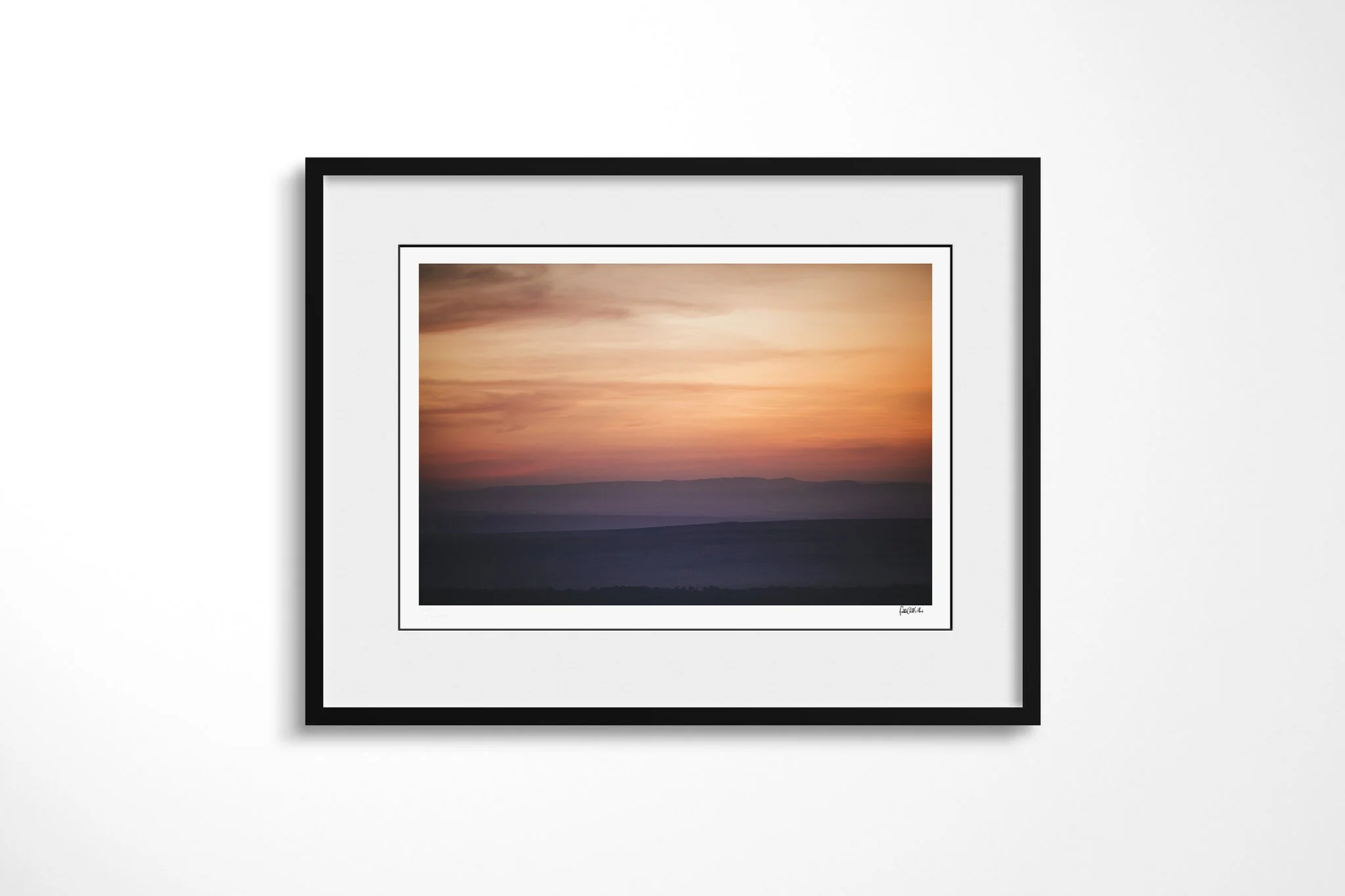 Framed fine art presentation of Sunrise in a black frame with a white mount, depicting soft horizontal bands of salmon, orange and mauve above blurred dark hills at sunrise.