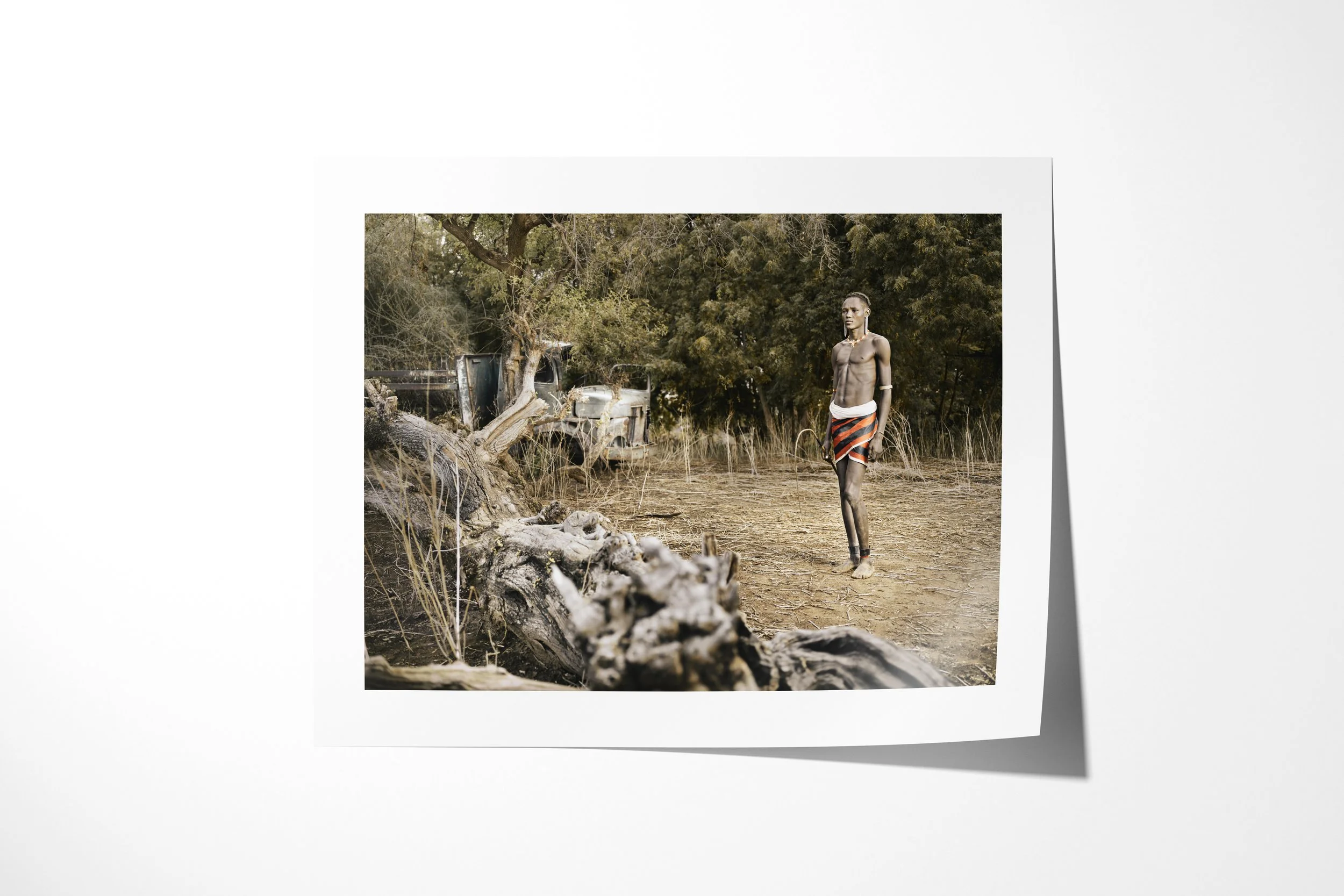 Fine art paper print of Lokoloi, showing a Daasanach man standing near an uprooted tree with an abandoned vehicle behind him.