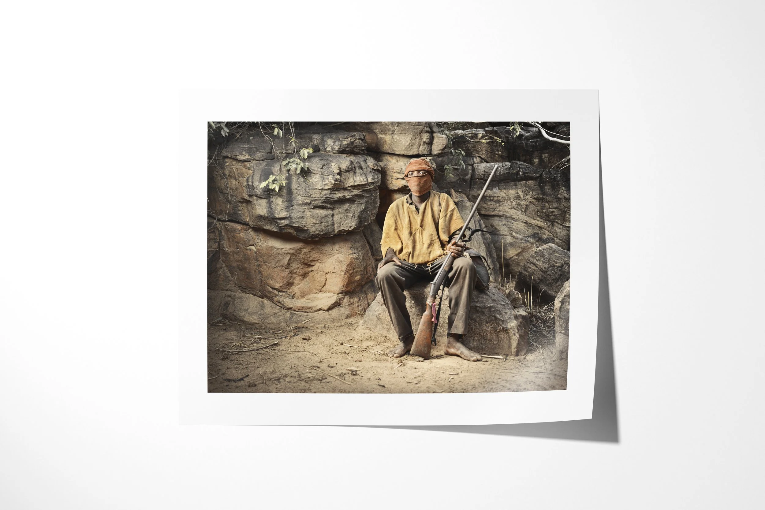 Fine art paper print of Duogolou, showing Duogolou seated barefoot against a rock face with his face wrapped and a rifle in hand.