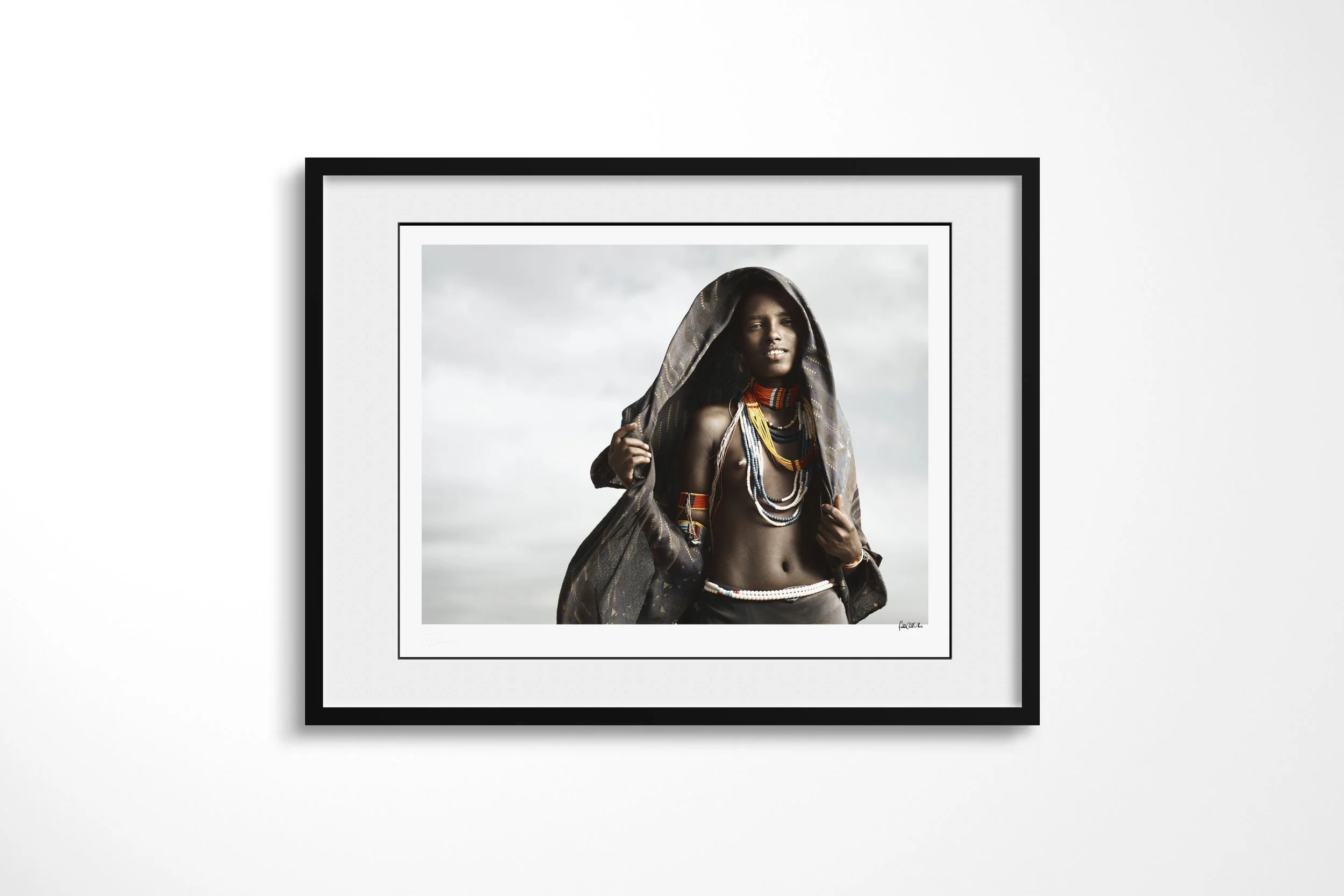 Framed presentation of Hara in a black frame and white mount, showing an unmarried Arbore woman wearing a dark head covering and layered coloured beadwork.