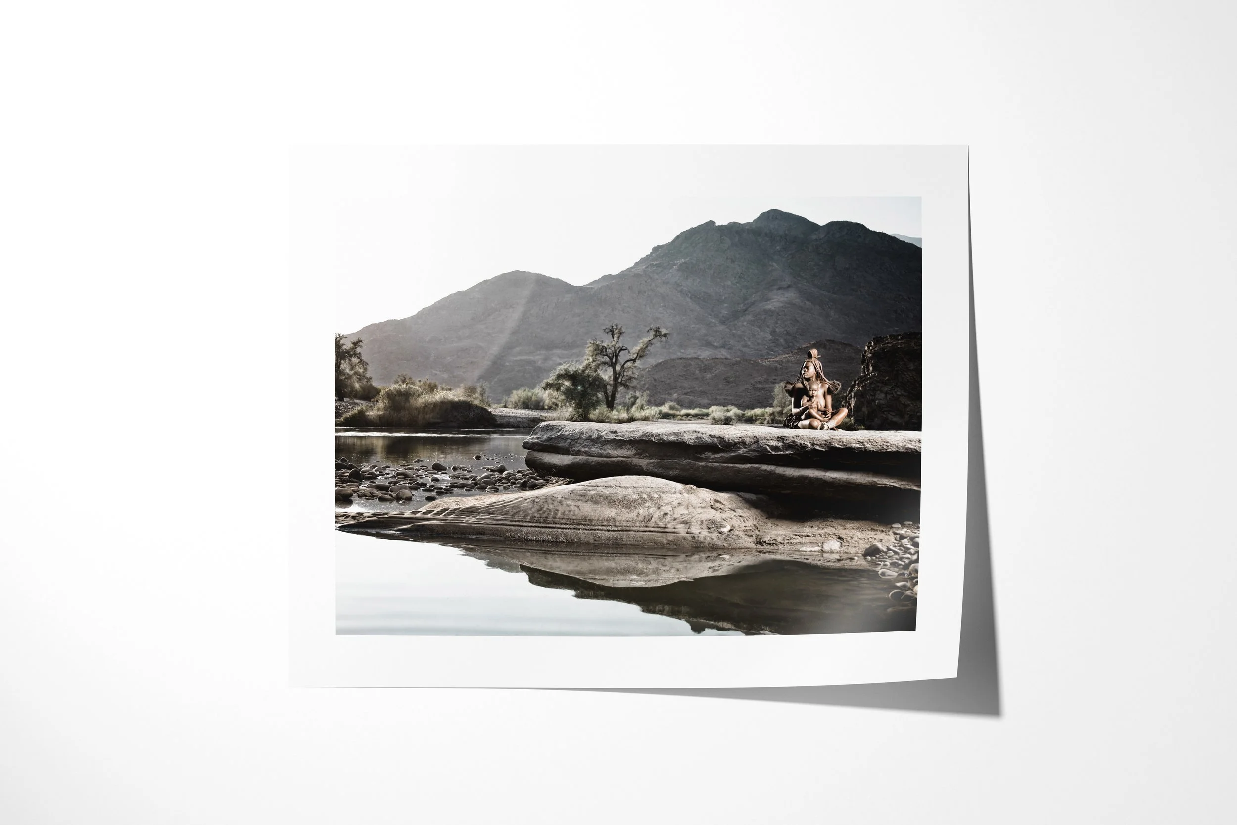 Fine art paper print of Tjikuria, showing Tjikuria and a child seated on a broad rock beside the Kunene River beneath dark mountains.