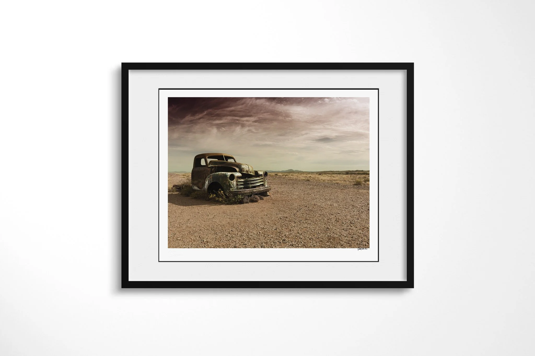 Framed presentation of Better Days in a black frame and white mount, showing a rusted vintage vehicle abandoned on a stony arid plain beneath dark, textured clouds and distant low mountains.